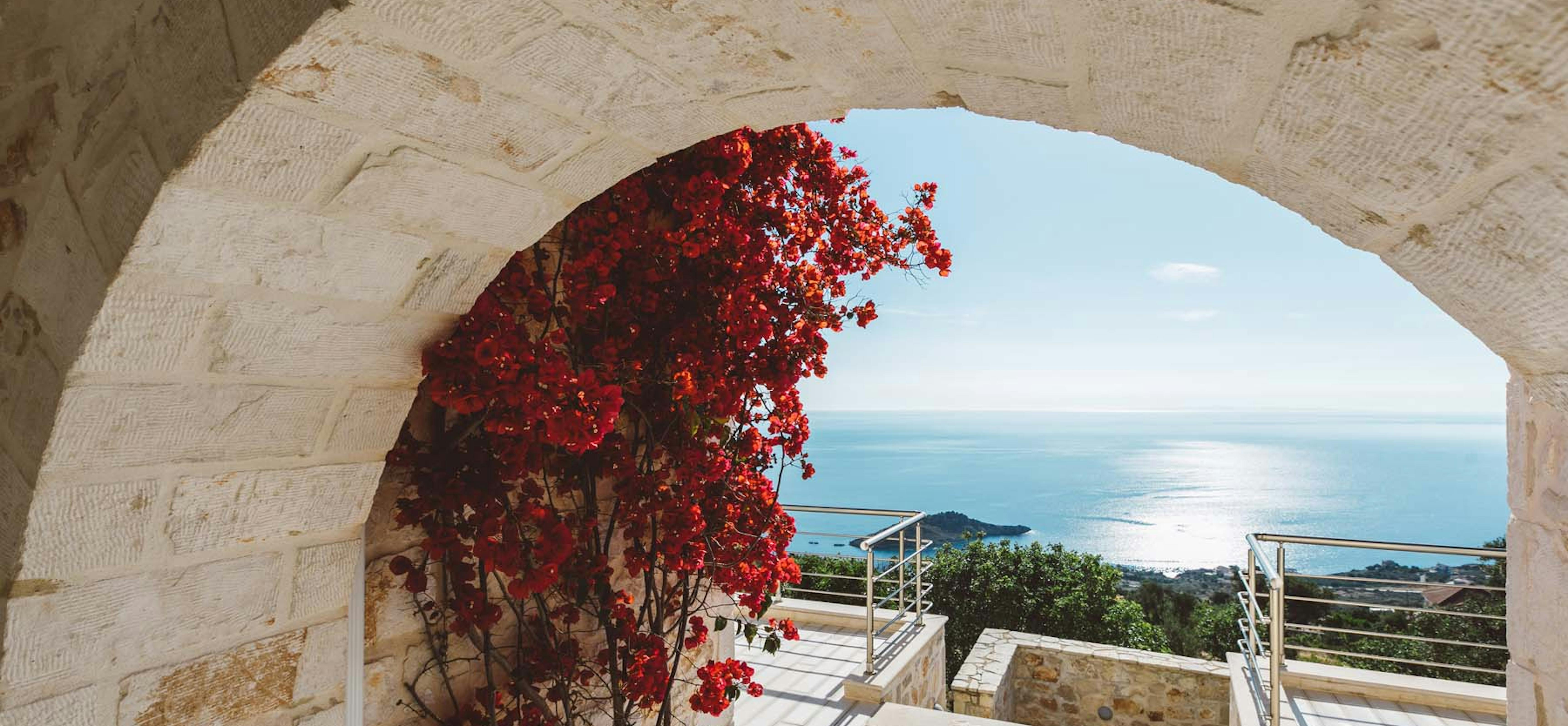 Stone archway framing terrace steps with vibrant red bougainvillea and ocean views beyond.
