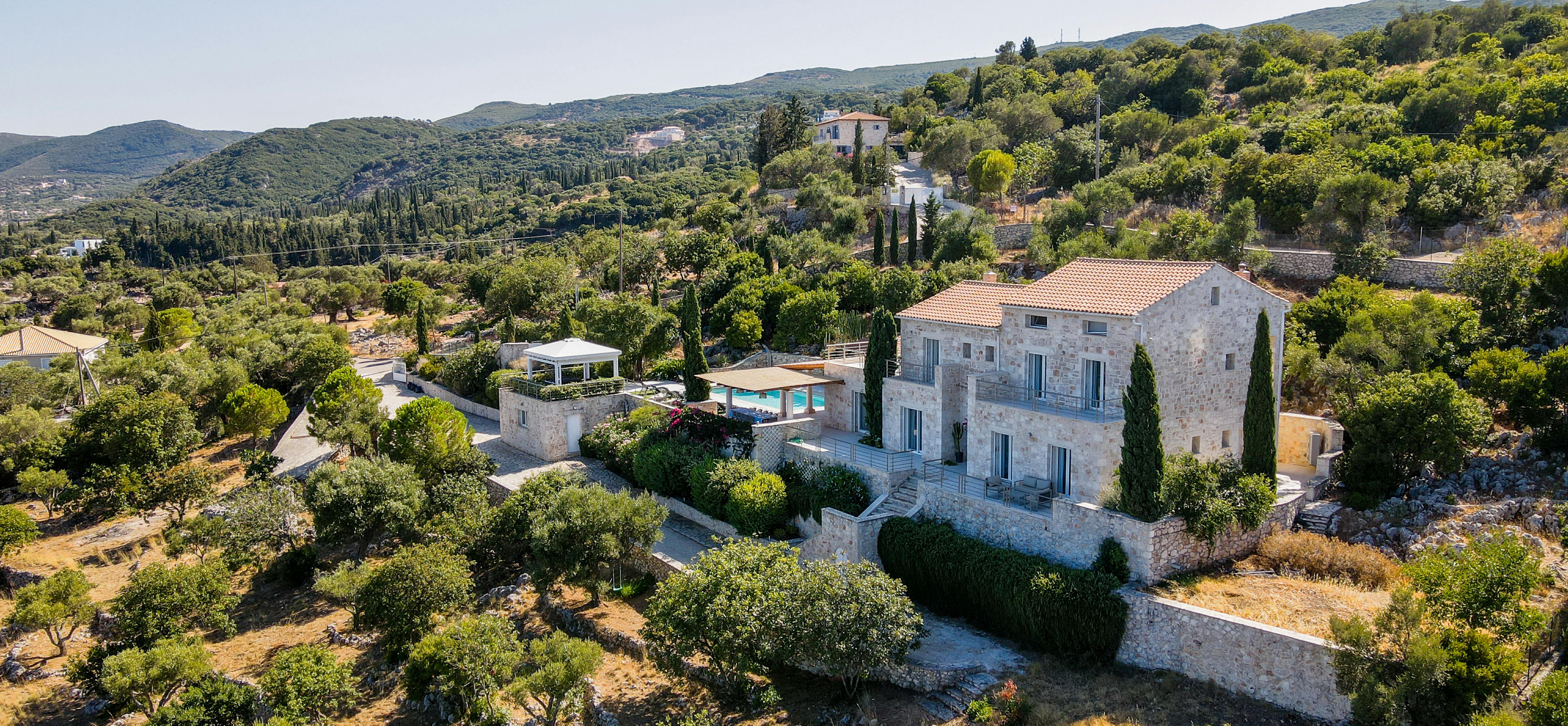 Aerial view of Mediterranean stone villa with terracotta roofs nestled in lush hillside landscape.