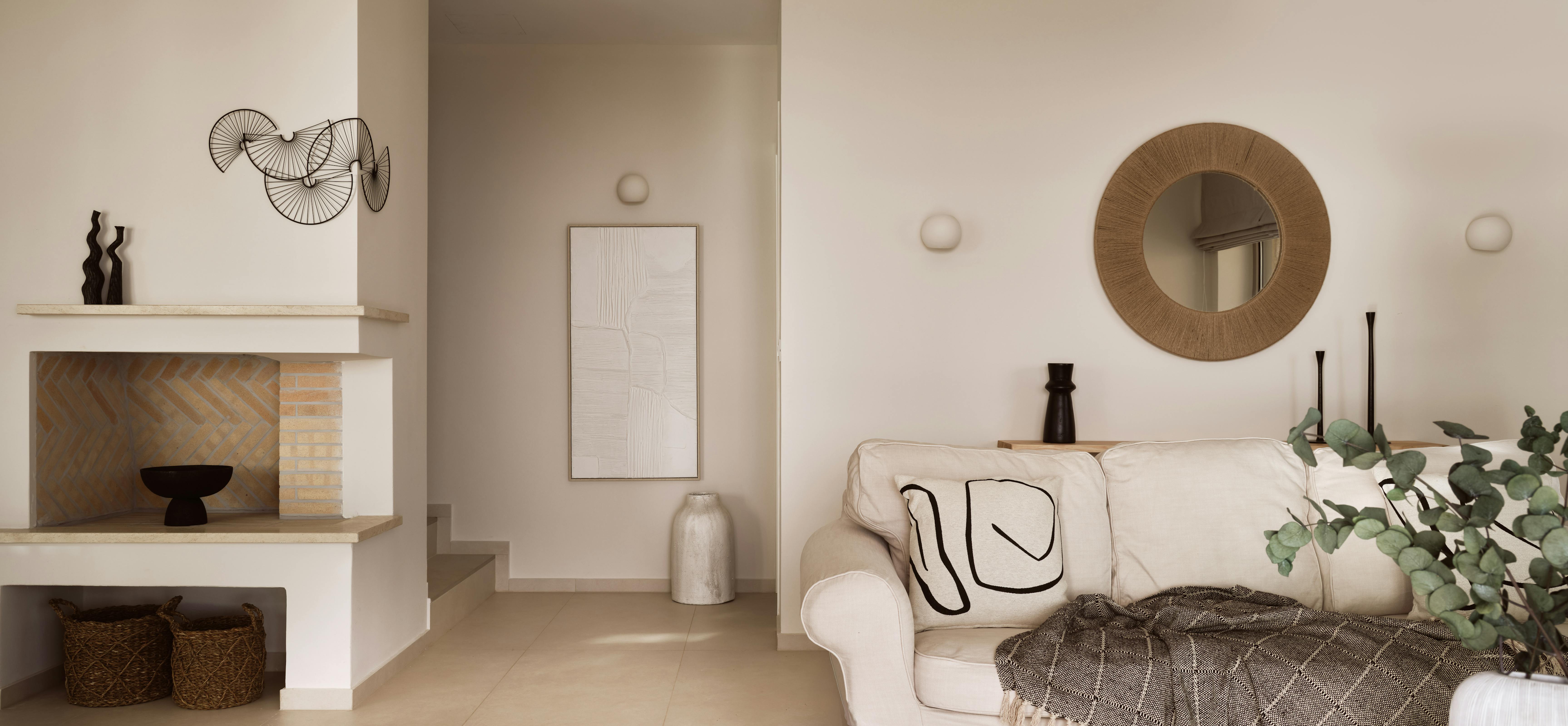 Contemporary living room with white fireplace, circular woven mirror, and minimalist decor in warm neutral palette.