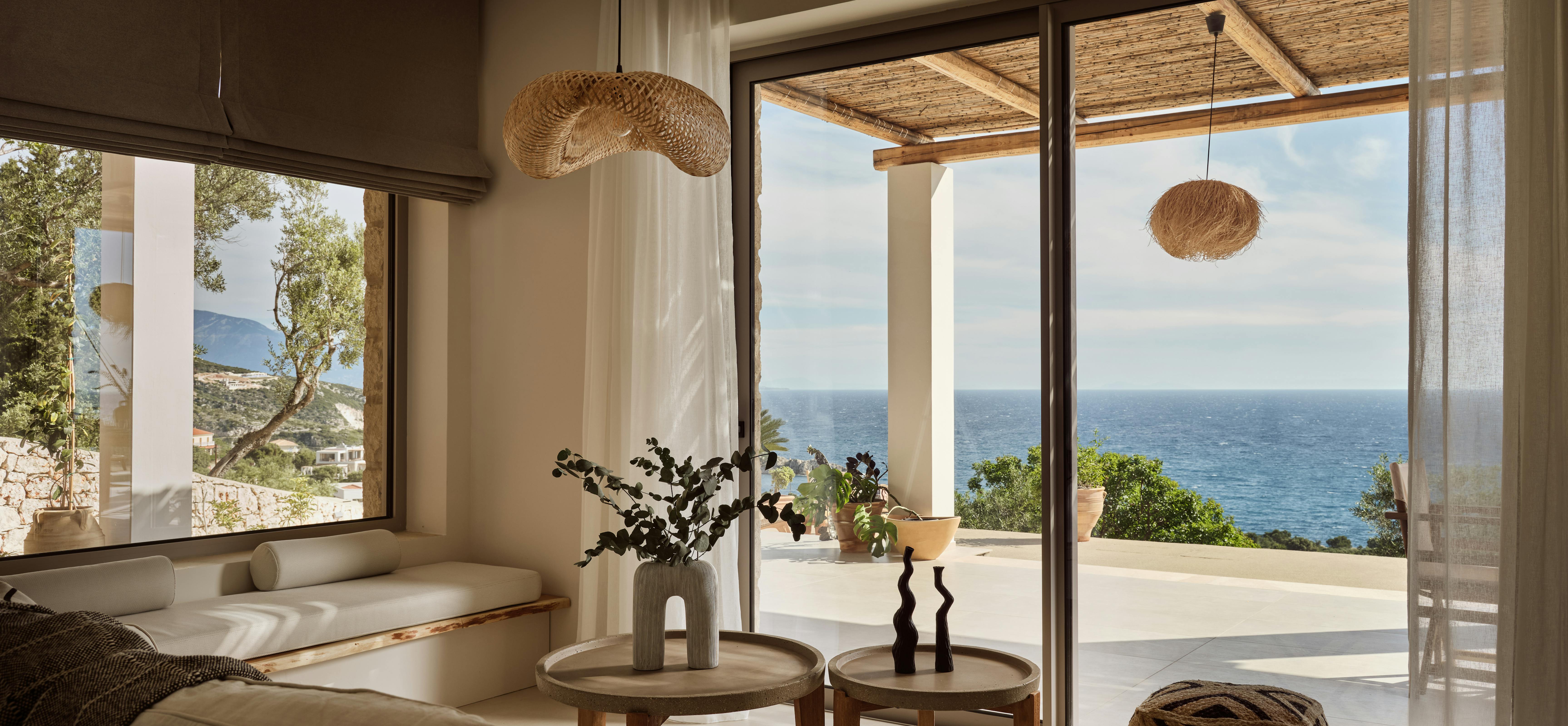Living area with built-in window seat, woven pendant lights, and panoramic doors framing Mediterranean coastal landscape.