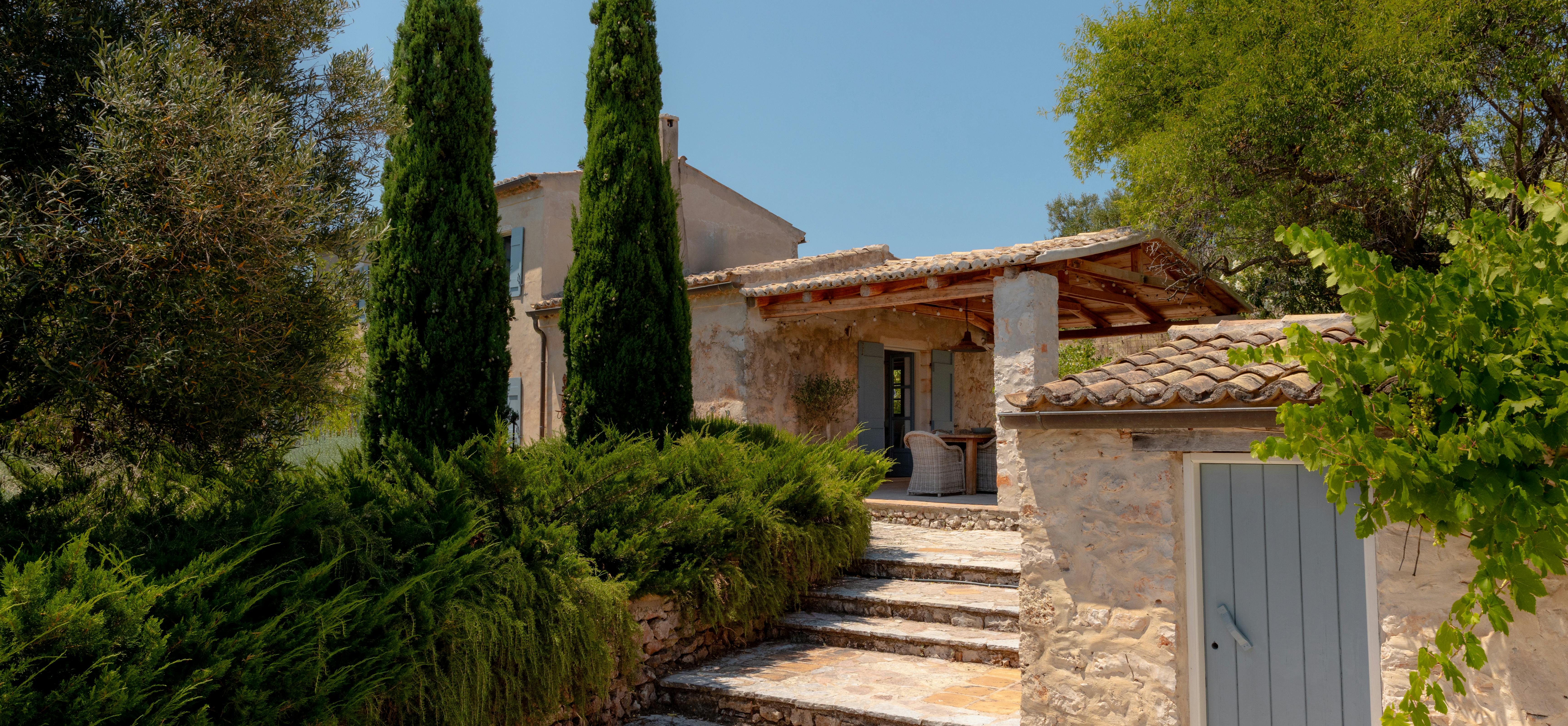 Stone villa entrance with curved steps flanked by cypress trees, leading to covered terrace with weathered walls and blue shutters.