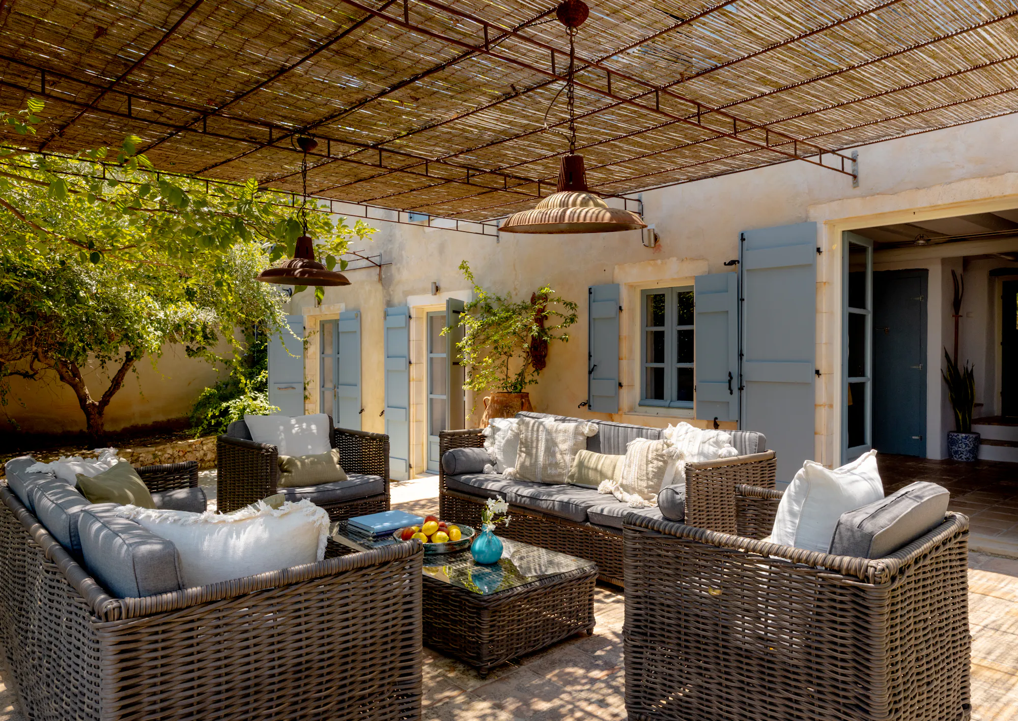 Covered terrace lounge with woven sofas under reed ceiling, facing stone facade with blue shutters and climbing vines.