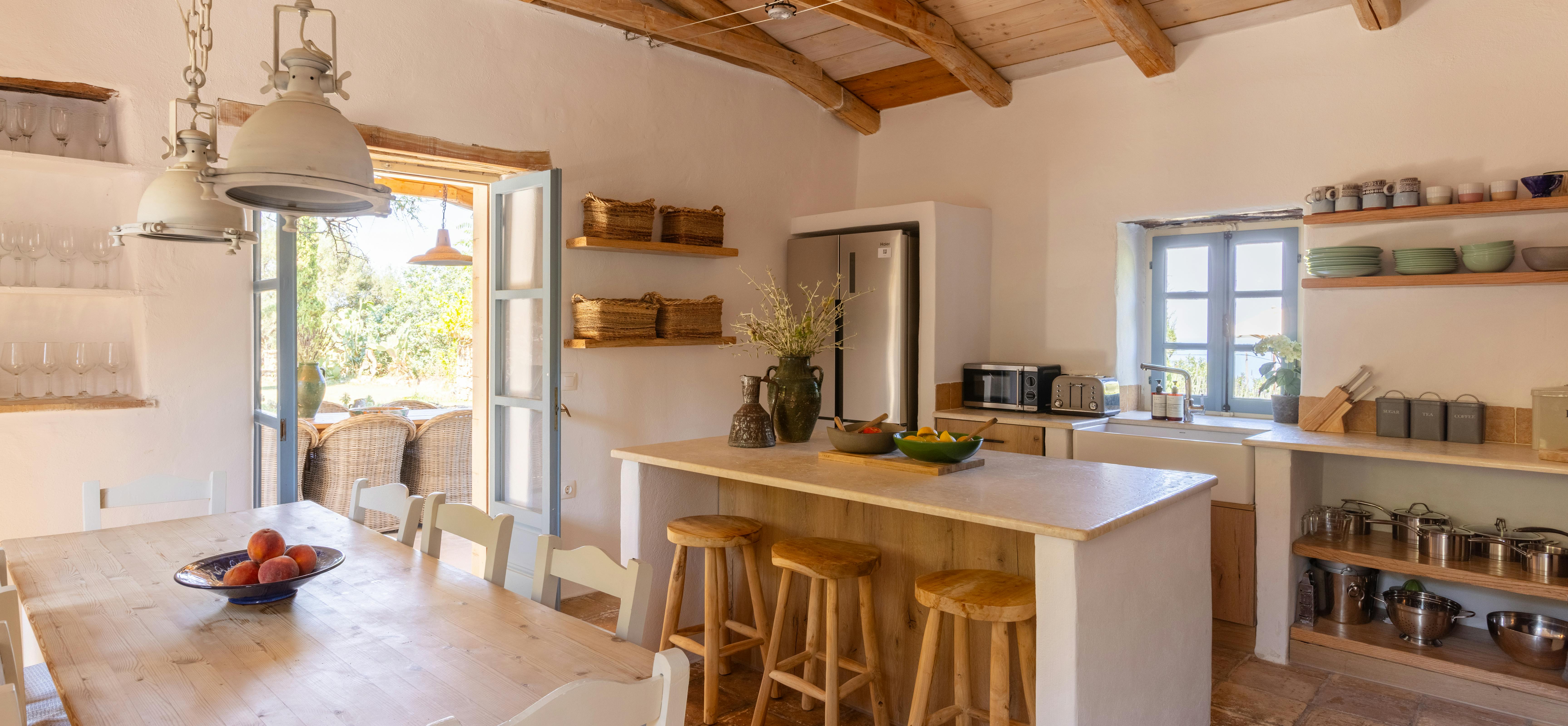Bright kitchen with wooden island, hanging pendant lights, open shelving, blue-shuttered windows, and rustic terracotta floor.