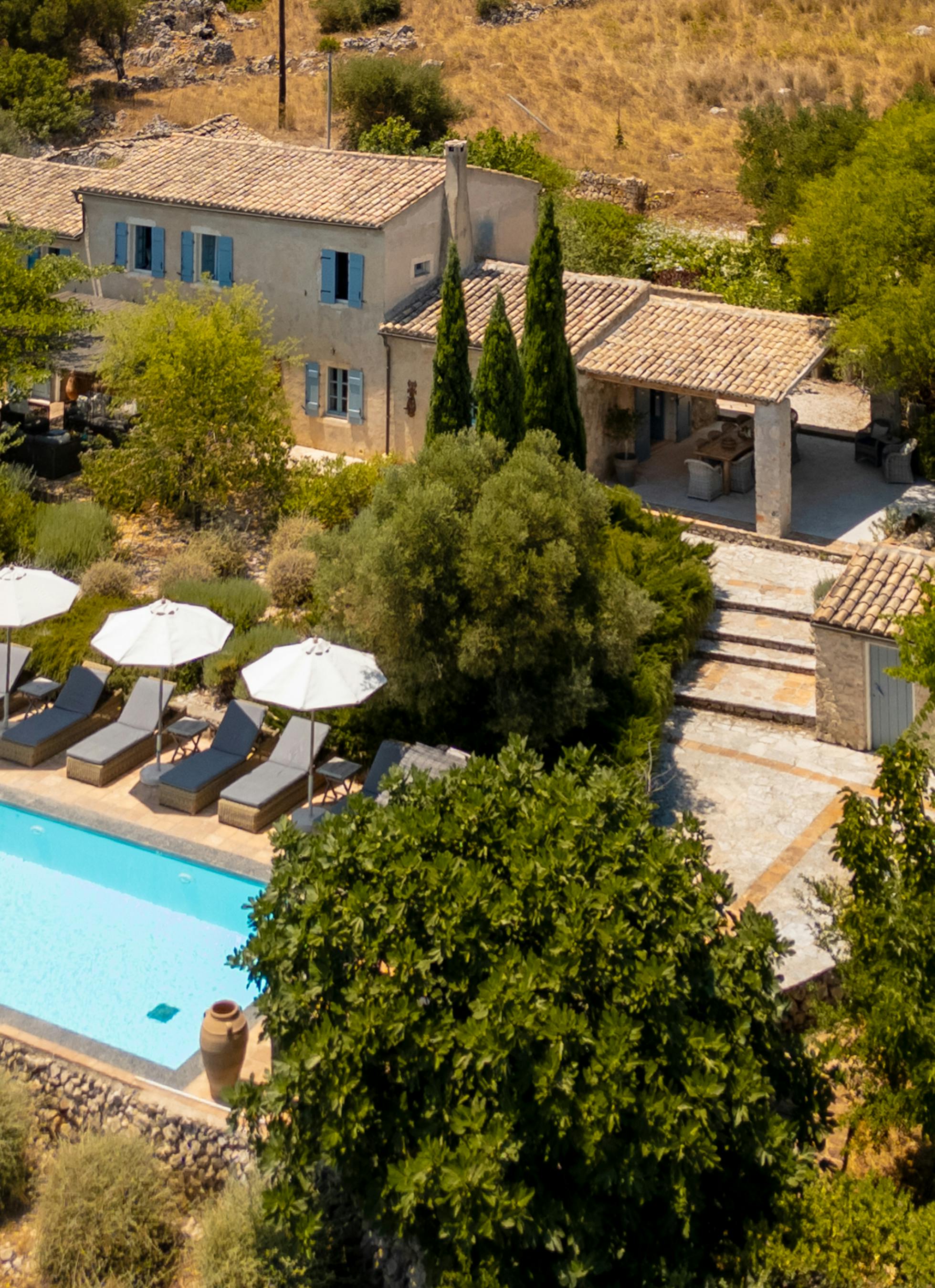 Aerial view of stone villa compound with rectangular pool, white umbrellas, and terraced gardens nestled in Mediterranean hillside.