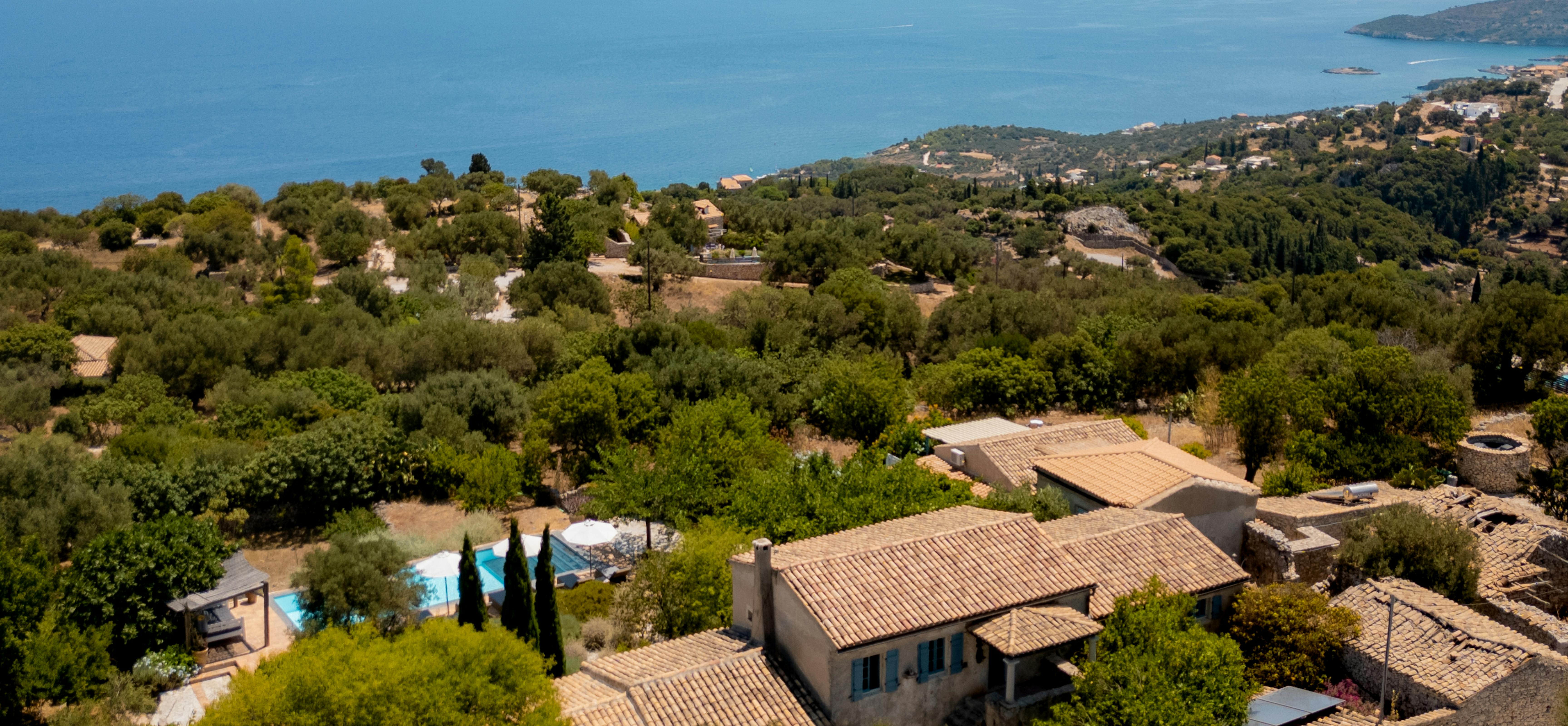 Aerial view of hillside villa with pool, terracotta roofs, cypress trees, and sweeping Mediterranean coastline beyond forested slopes.