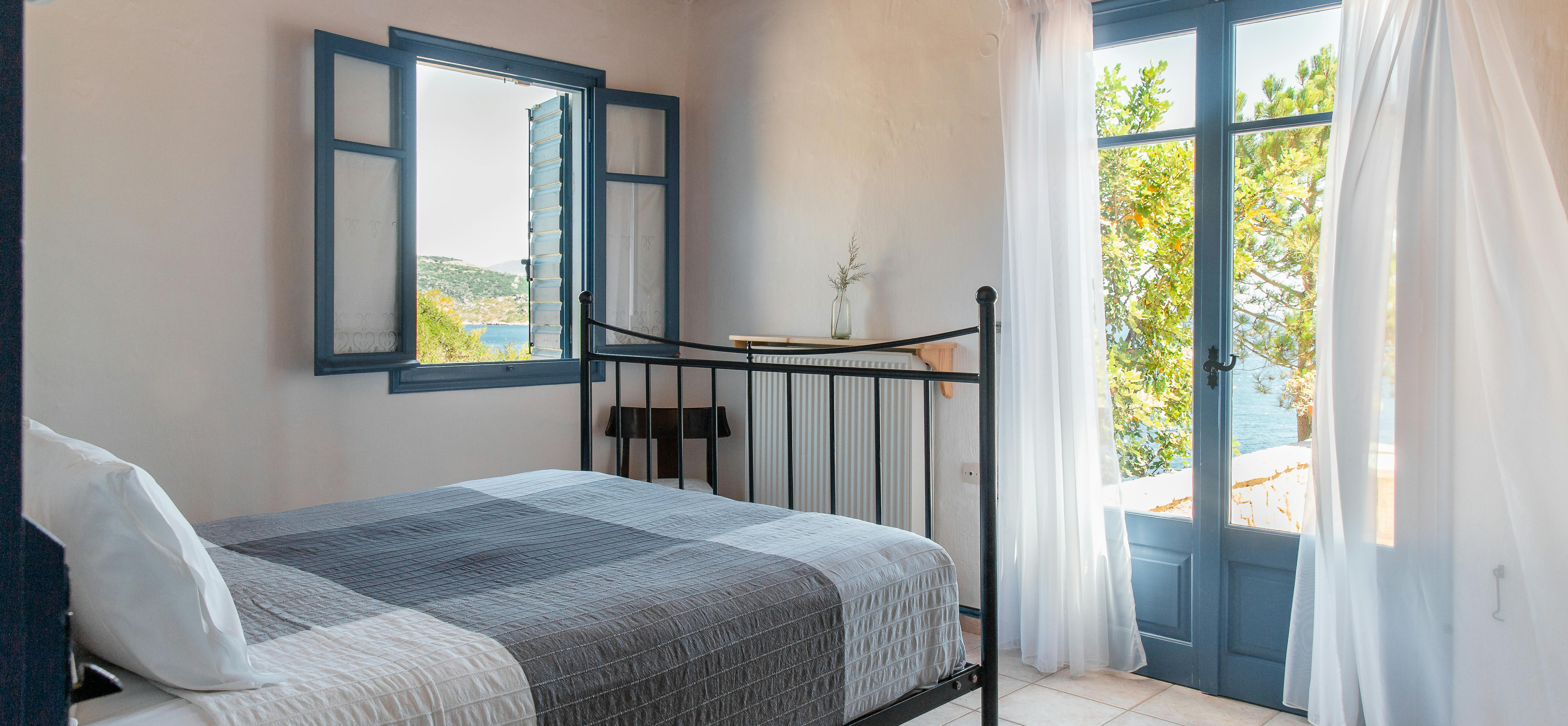Airy bedroom with metal bed frame, exposed wood beams, blue-trimmed windows and door opening to garden terrace.