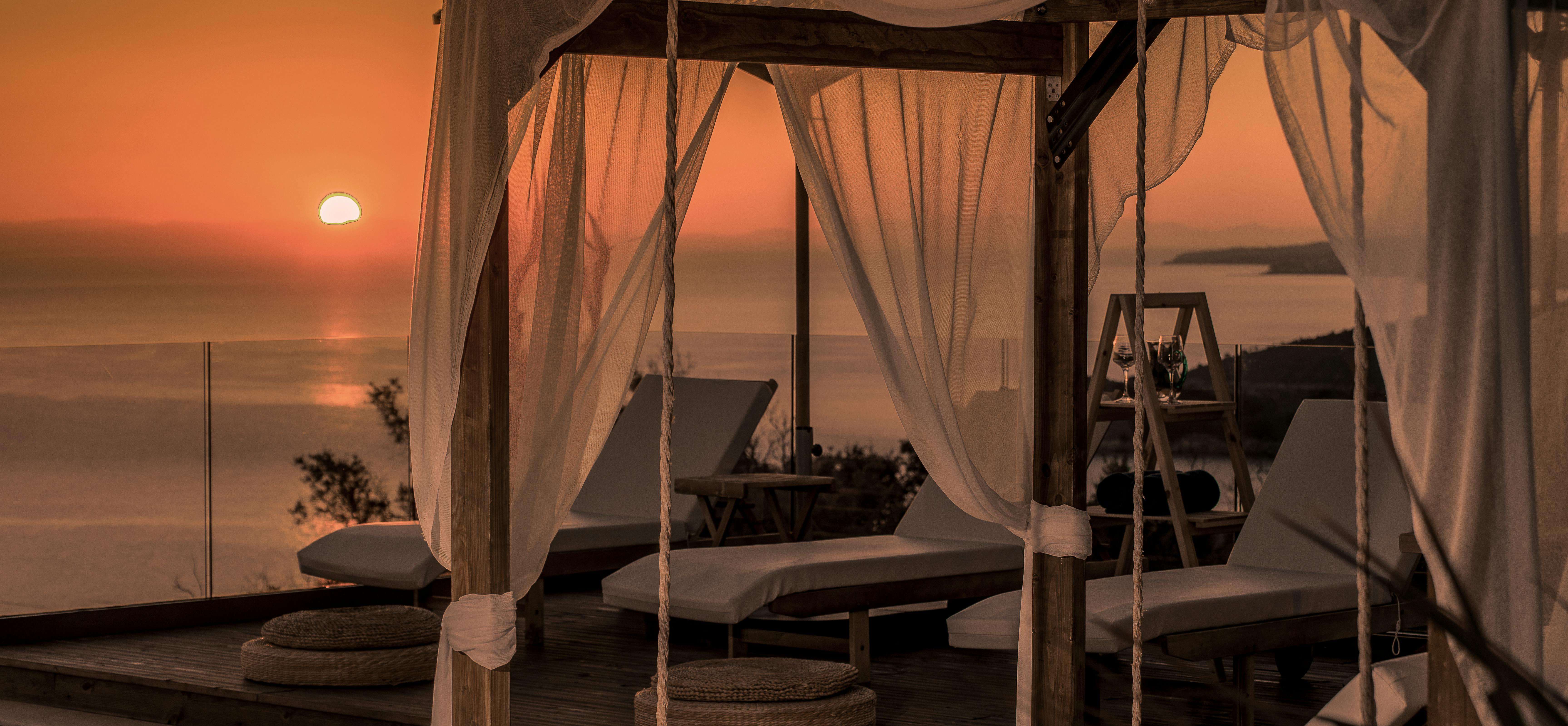 Dusk poolside scene with draped wooden cabanas, sheer curtains billowing in breeze, and fiery orange sunset over calm sea.