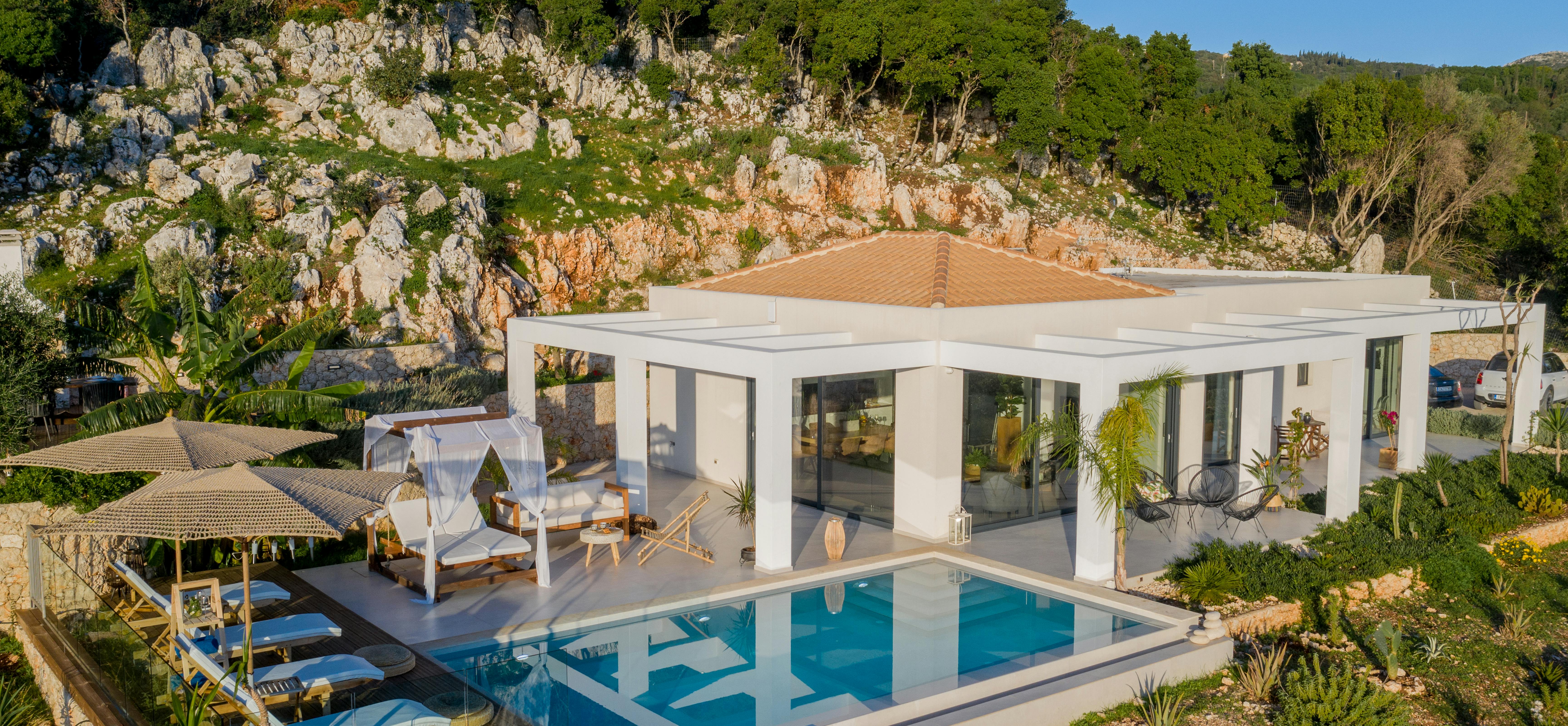 Aerial view of minimalist white villa with infinity pool, canopy daybed, and thatched pavilion nestled against dramatic limestone cliff.