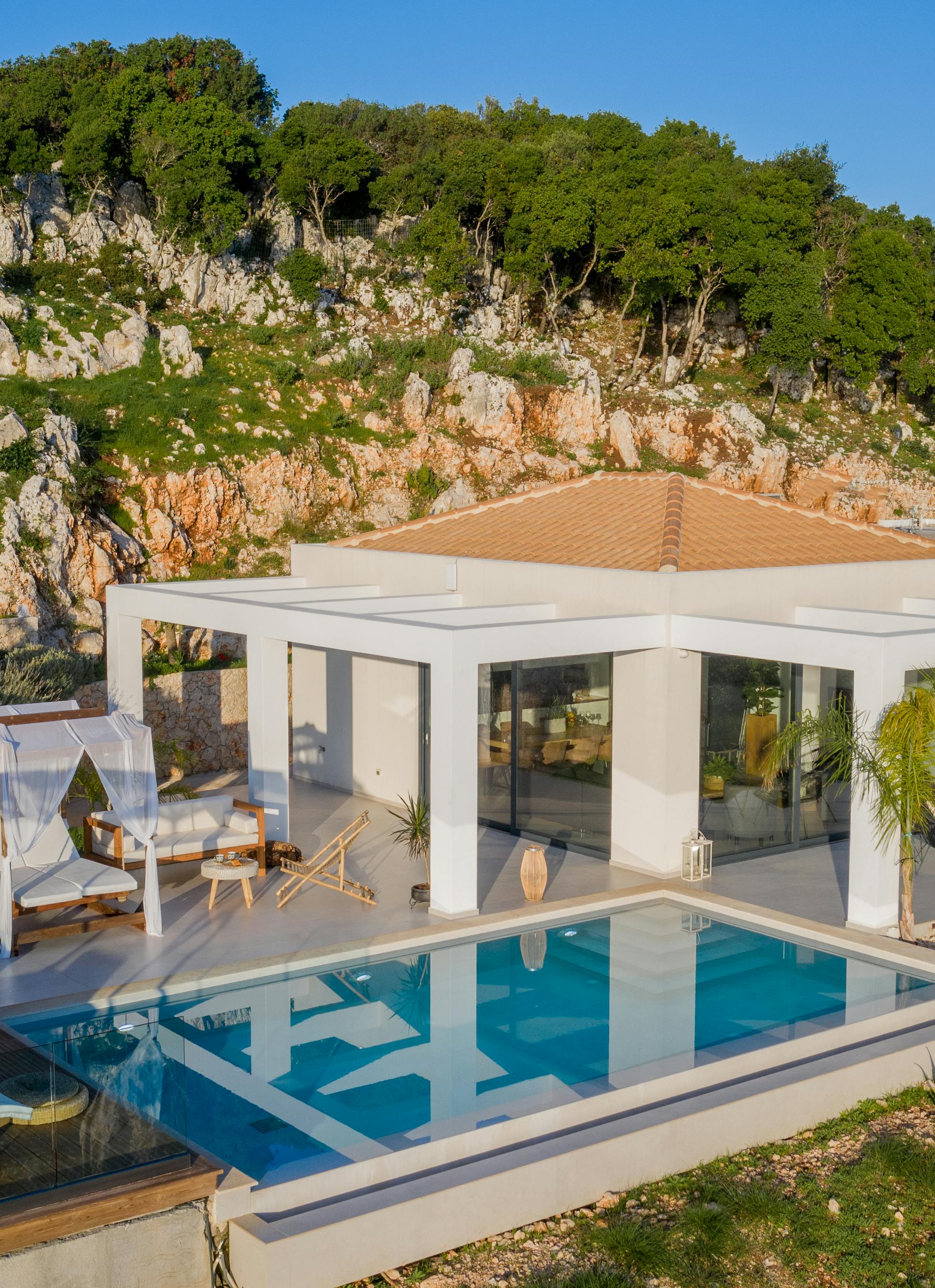 Aerial view of minimalist white villa with infinity pool, canopy daybed, and thatched pavilion nestled against dramatic limestone cliff.