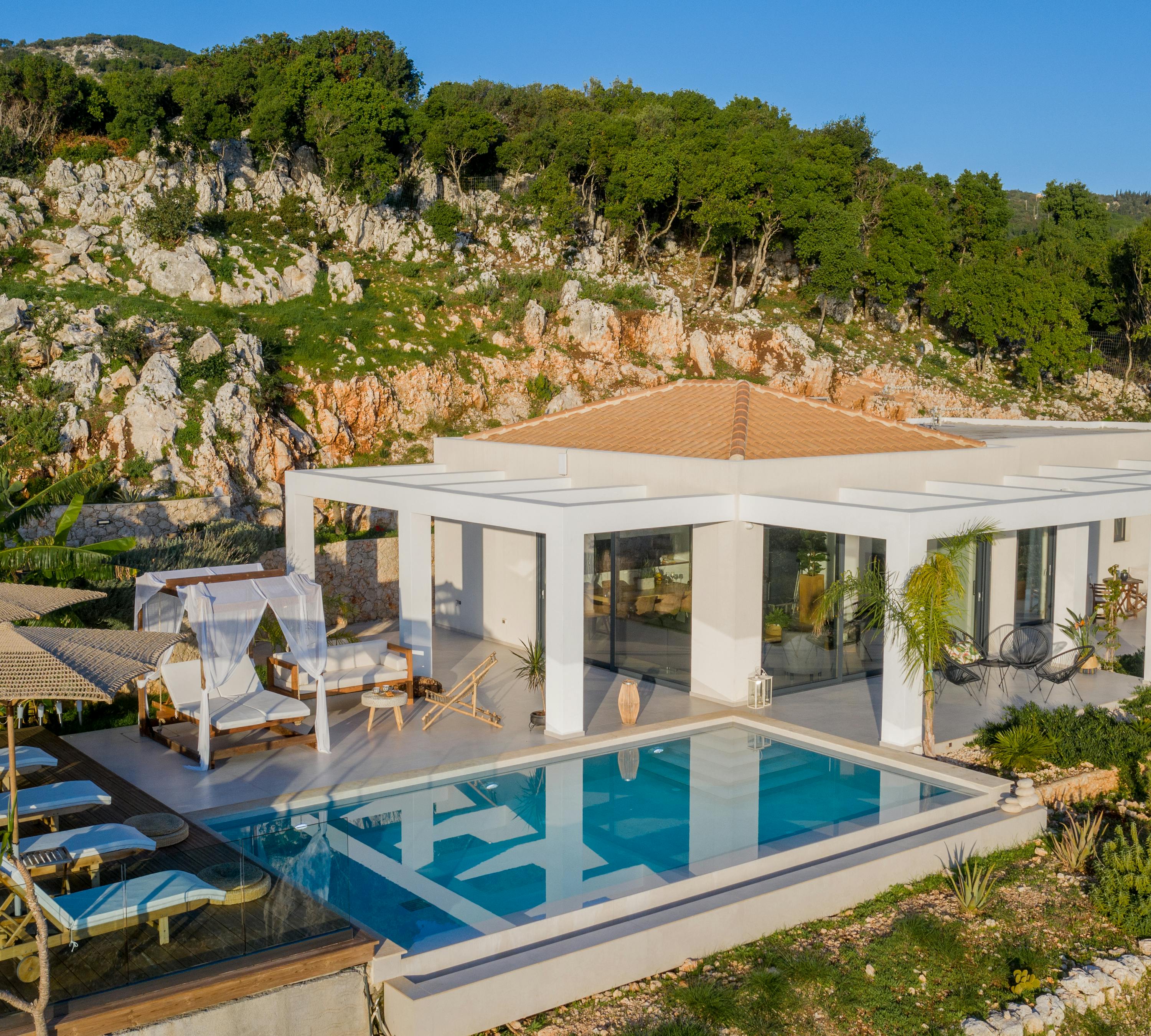 Aerial view of minimalist white villa with infinity pool, canopy daybed, and thatched pavilion nestled against dramatic limestone cliff.