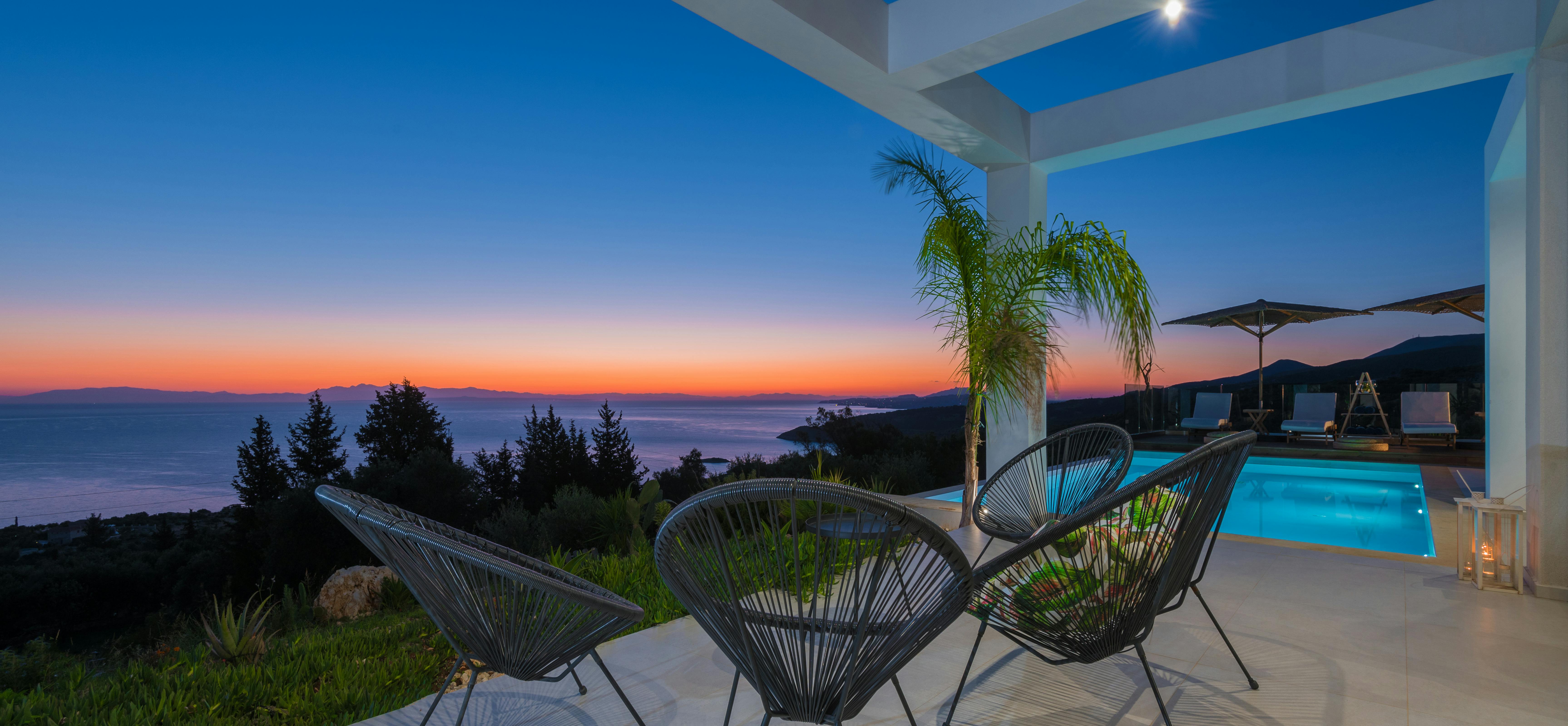Twilight terrace with modern wire chairs under white pergola, overlooking illuminated infinity pool and coastal sunset gradient sky.