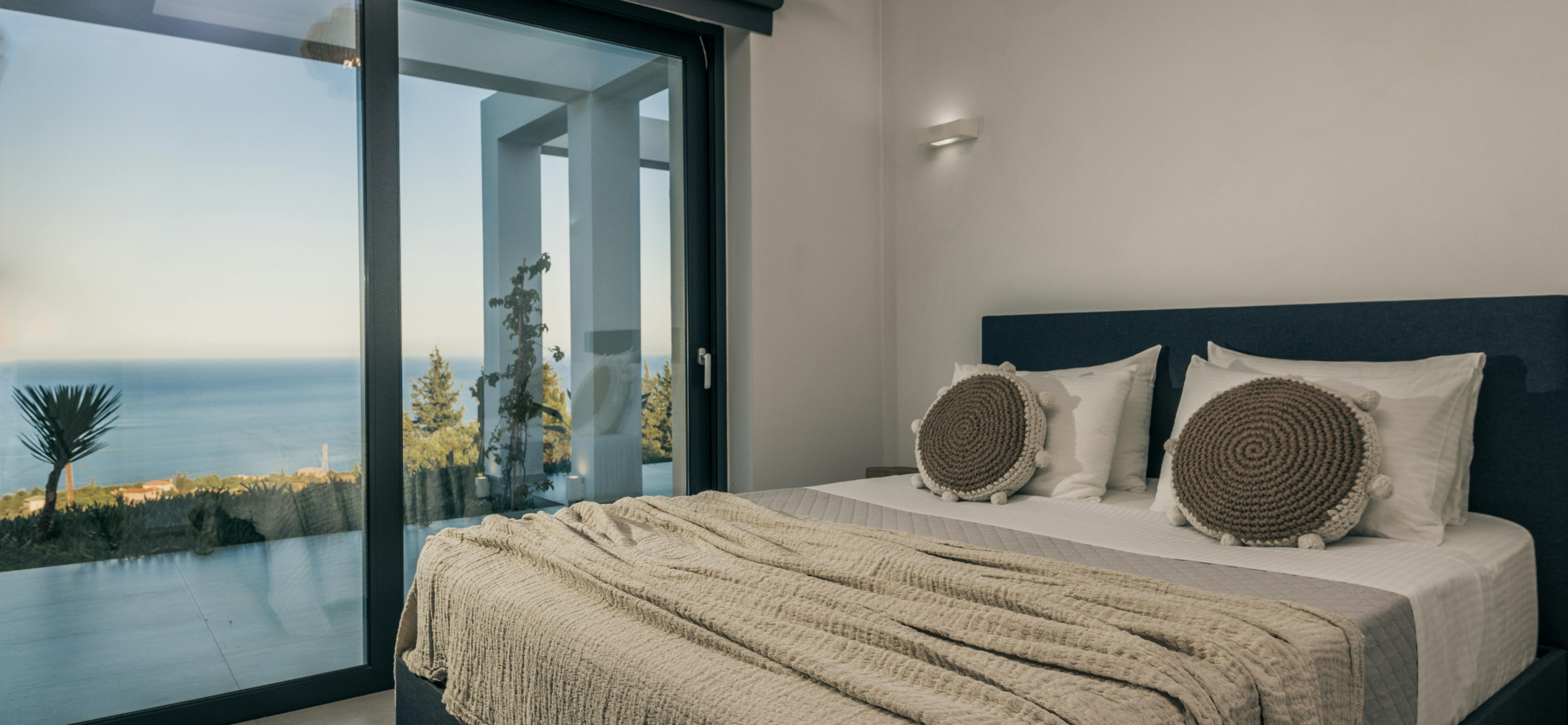 Minimalist bedroom with dark upholstered bed, woven circular pillows, and sliding glass doors opening to infinity pool with sea views.