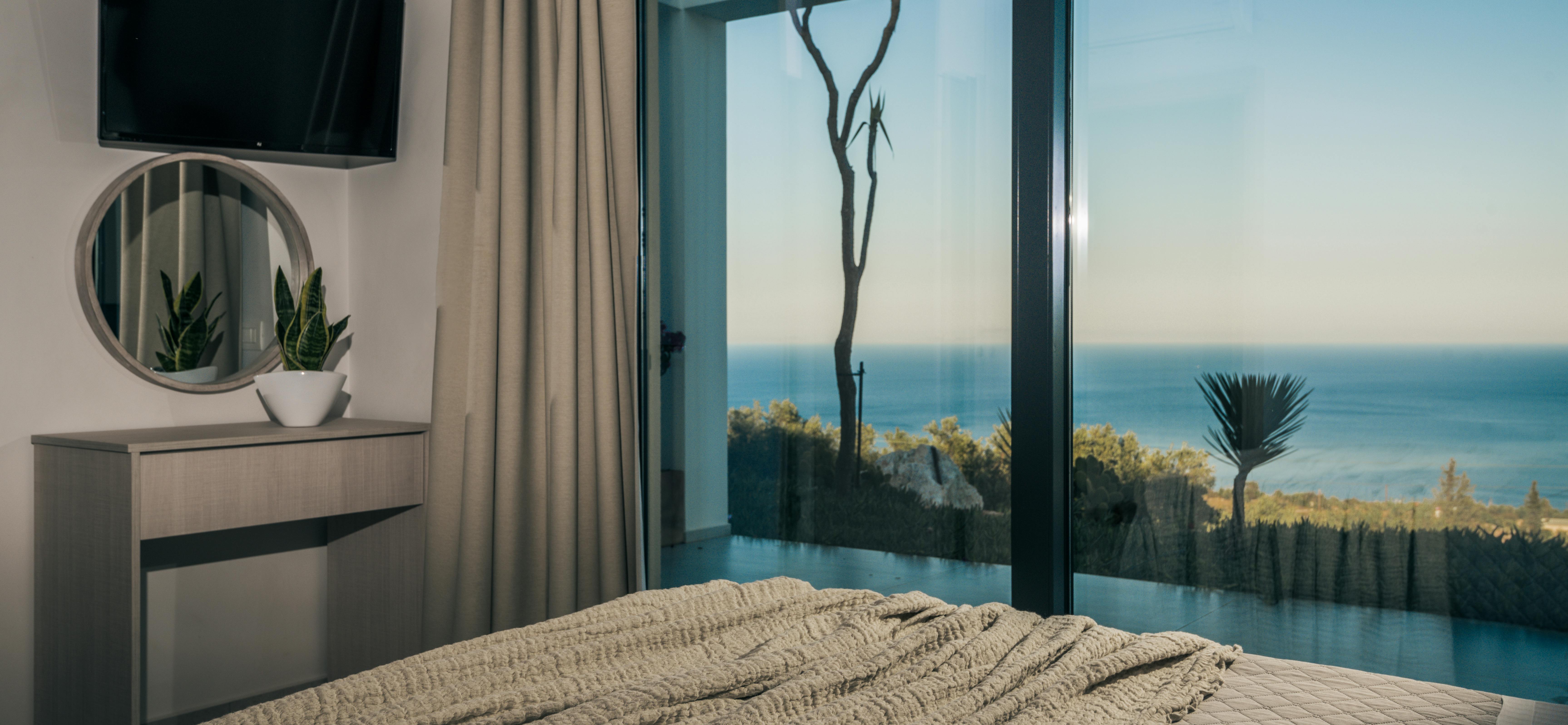 Serene bedroom with textured bedding, round mirror, low dresser, and floor-to-ceiling windows framing infinity pool and ocean beyond.