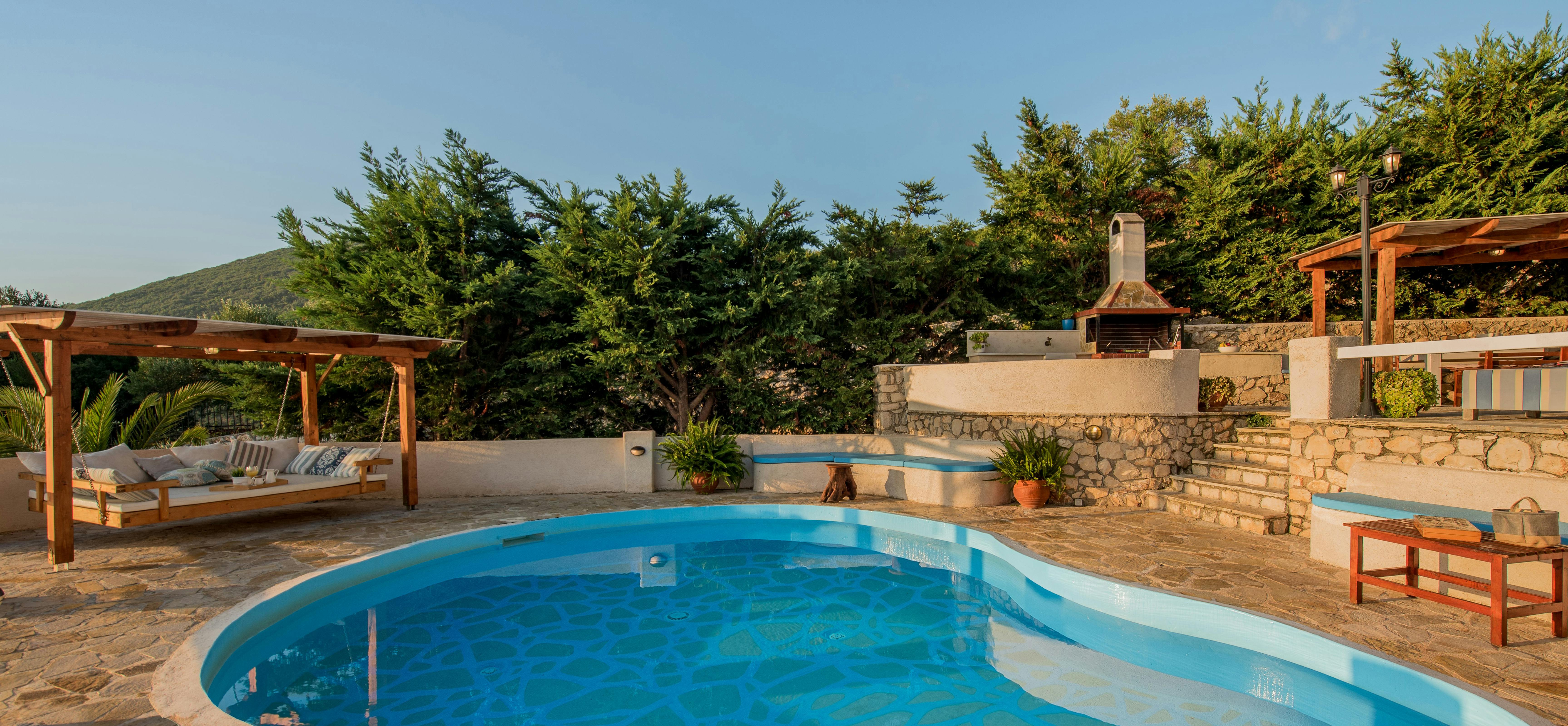 Curved infinity pool with stone decking, wooden pergola daybed, and terraced levels featuring outdoor grill and mountain backdrop.
