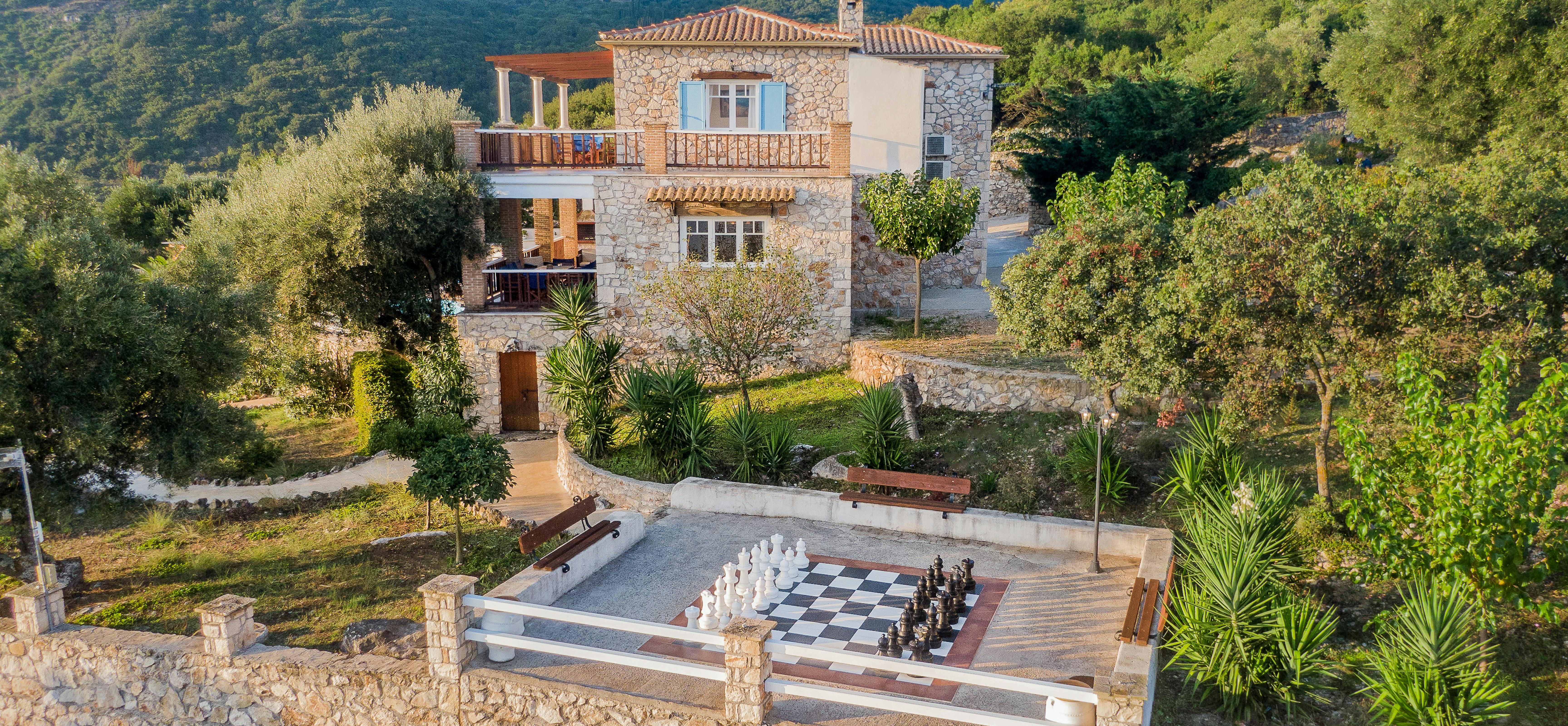 Aerial view of terraced stone villa with giant chess board patio, surrounded by Mediterranean gardens and mountain landscape.