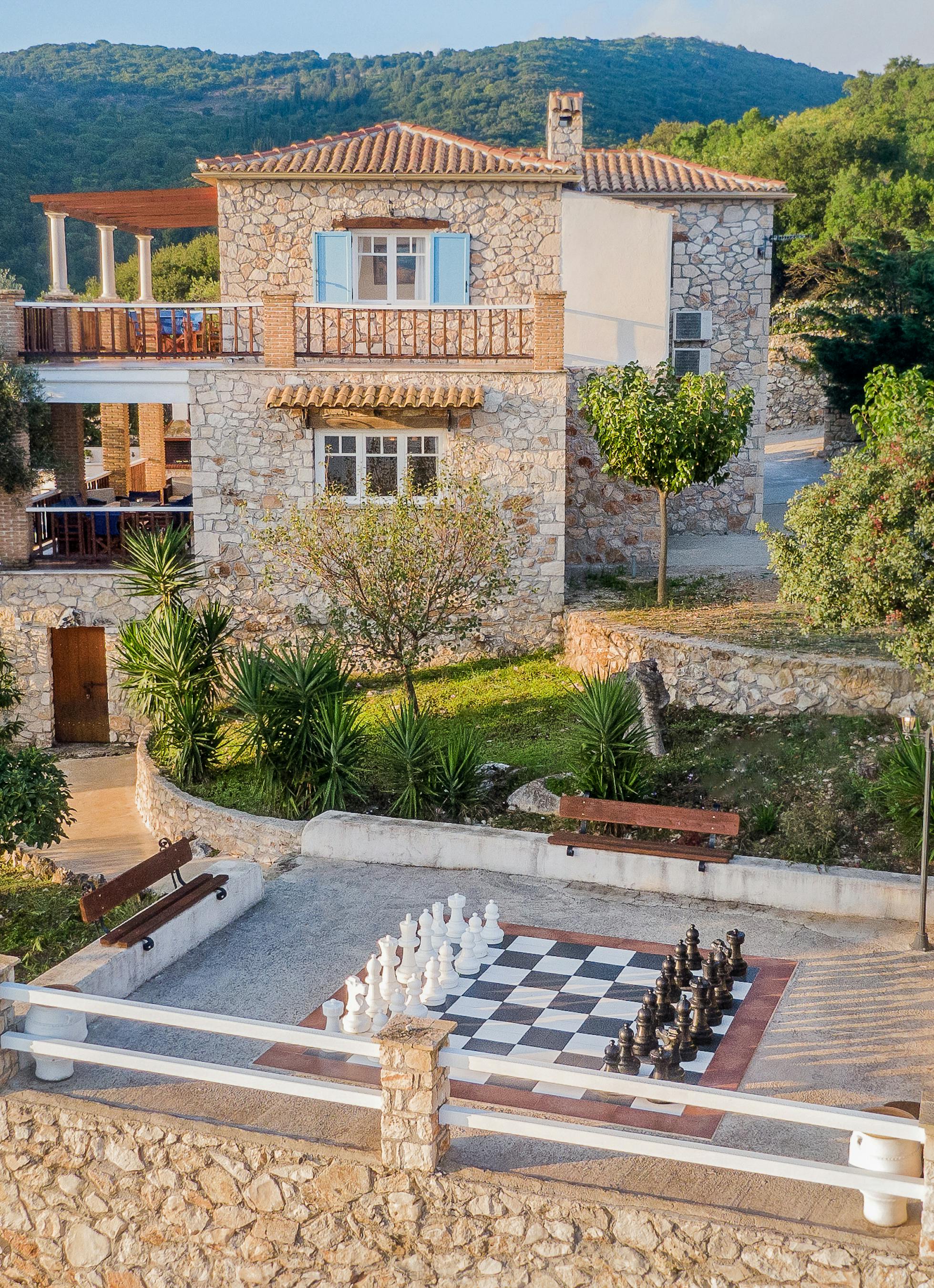 Aerial view of terraced stone villa with giant chess board patio, surrounded by Mediterranean gardens and mountain landscape.