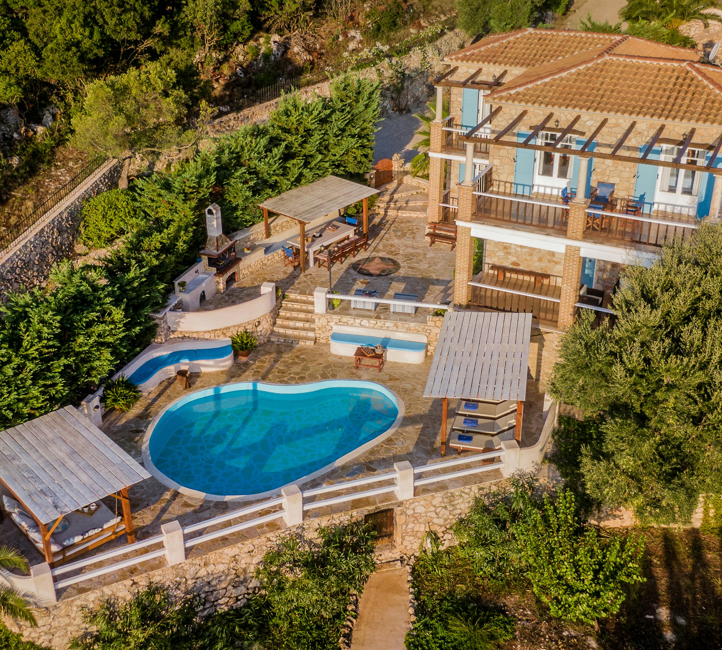 Aerial perspective of hillside villa with curved pool, multiple wooden pergolas, terraced levels, and olive grove surroundings.