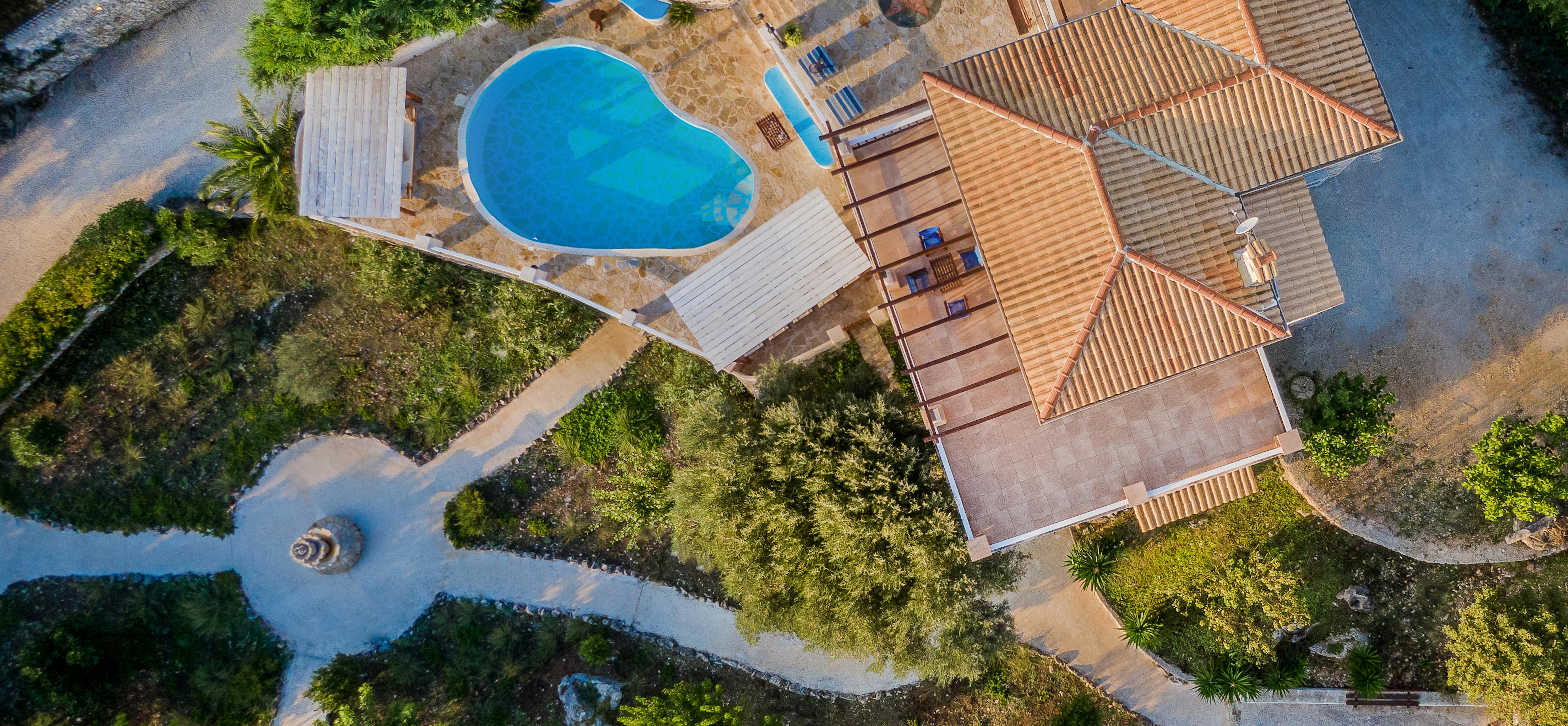 Aerial view showing organic-shaped pool, terracotta villa roofs, wooden decking, and winding pathways through lush gardens.