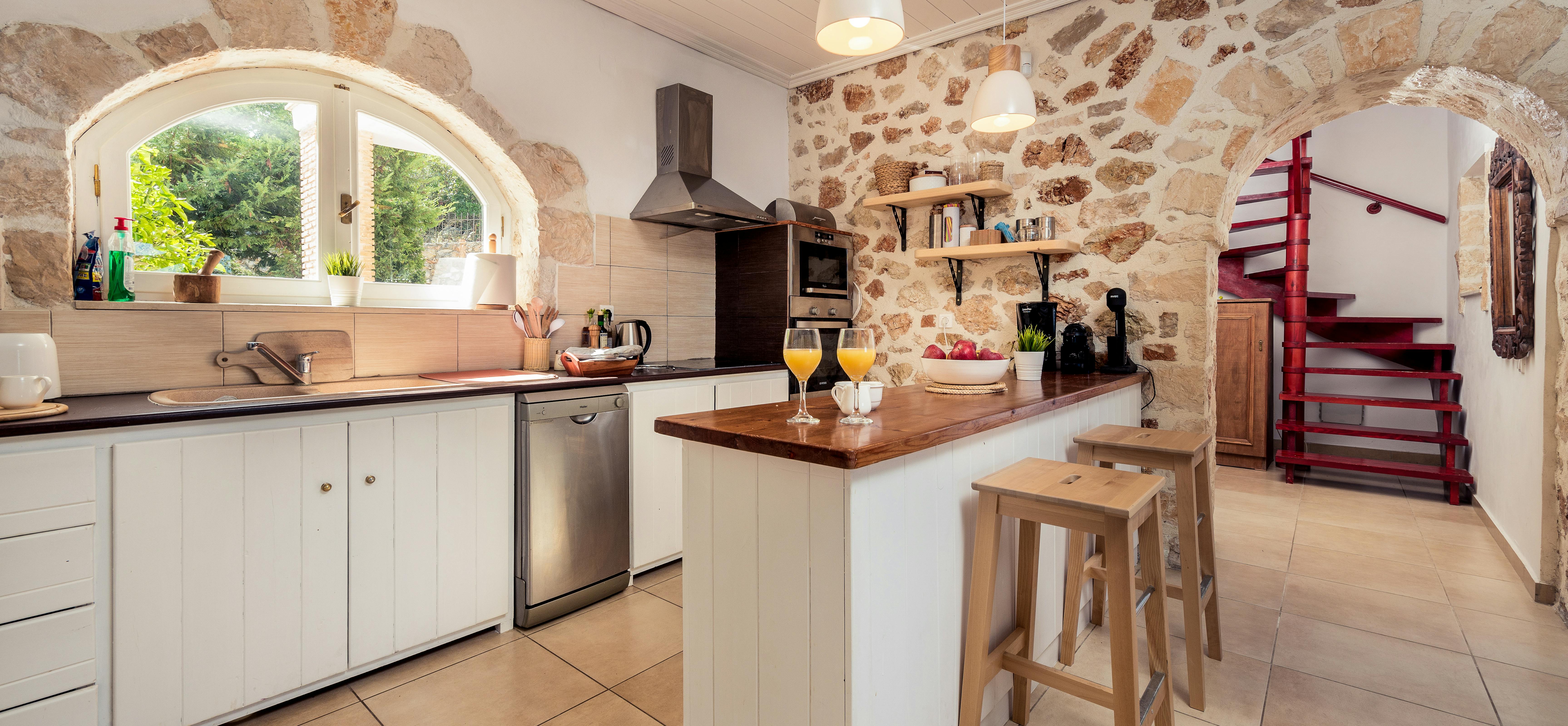 Rustic kitchen with exposed stone walls, white shaker cabinets, arched garden window, and wooden breakfast bar with stools.
