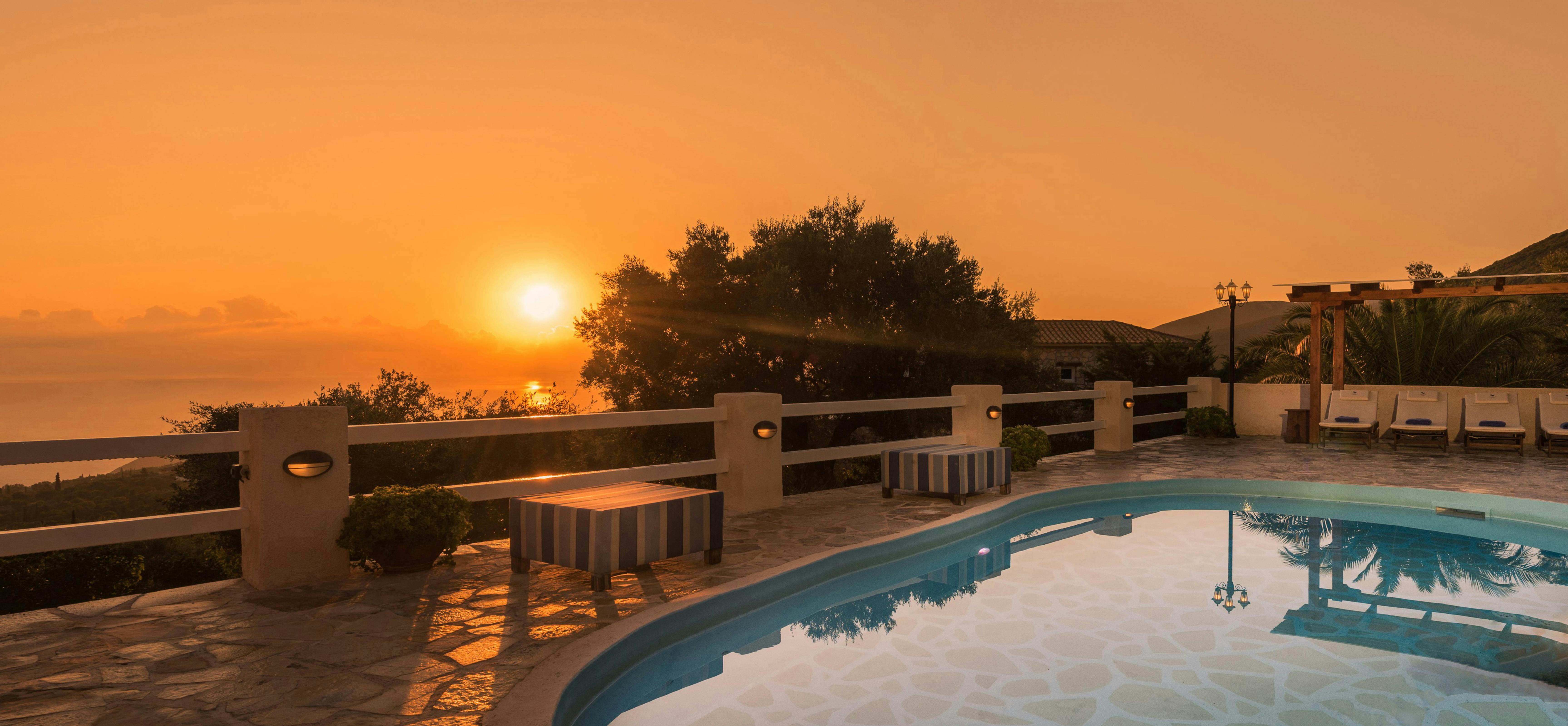 A curved swimming pool at sunset overlooks the sea, with a stone-topped bench nearby and warm orange light illuminating the terrace and distant hills.