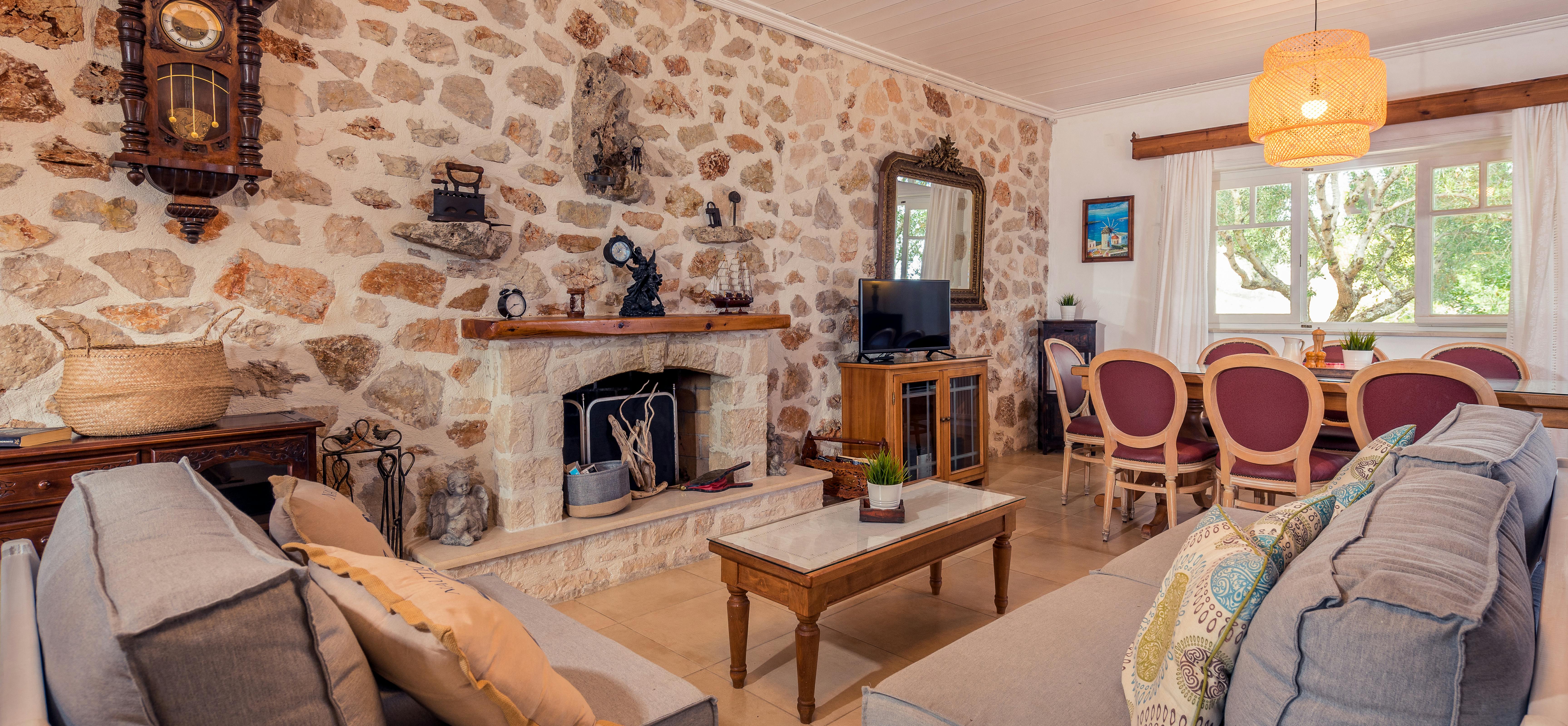 A living room with exposed stone walls centres on a fireplace adorned with vintage items, flanked by grey sofas and a dining area with burgundy chairs.