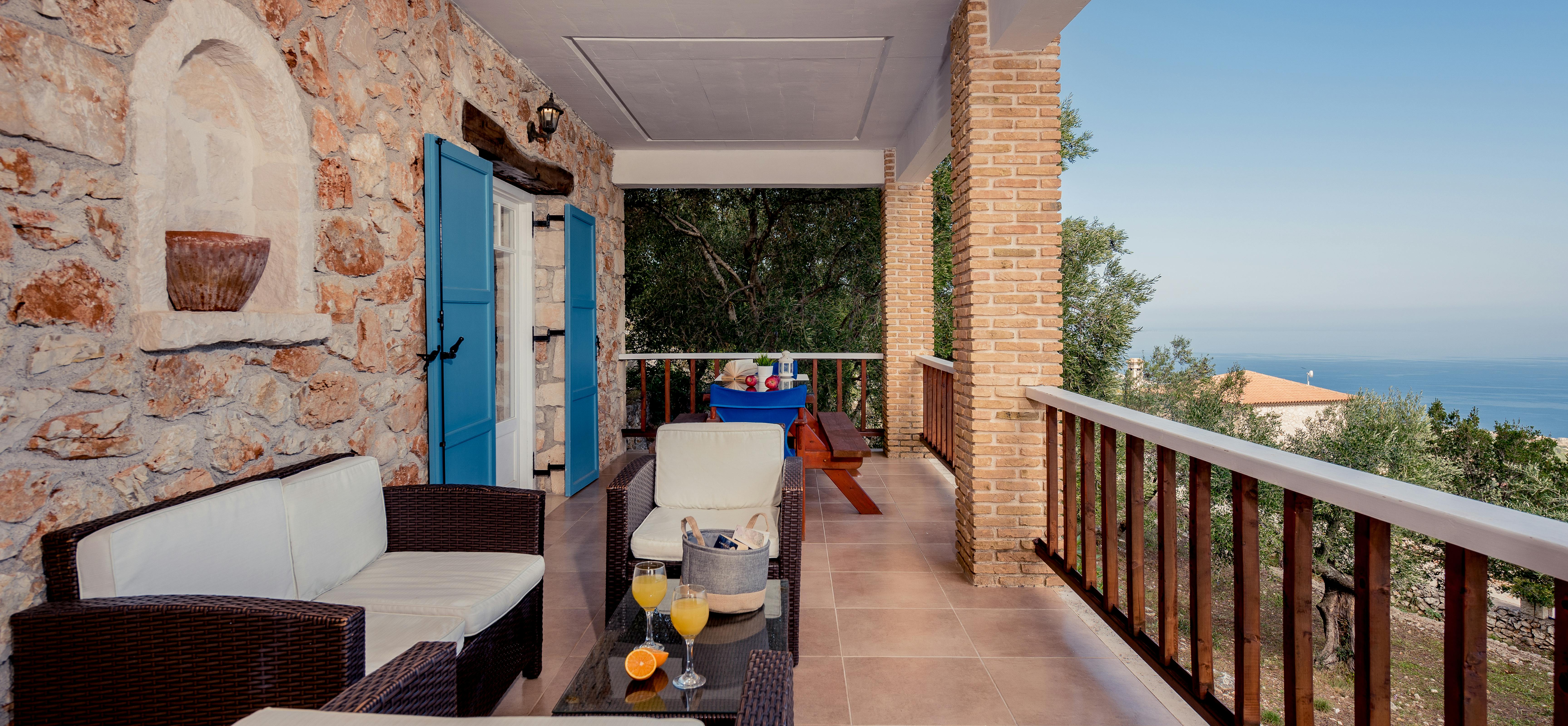 A covered balcony furnished with wicker seating and blue cushions offers panoramic sea views, with exposed stone walls and a blue door visible.