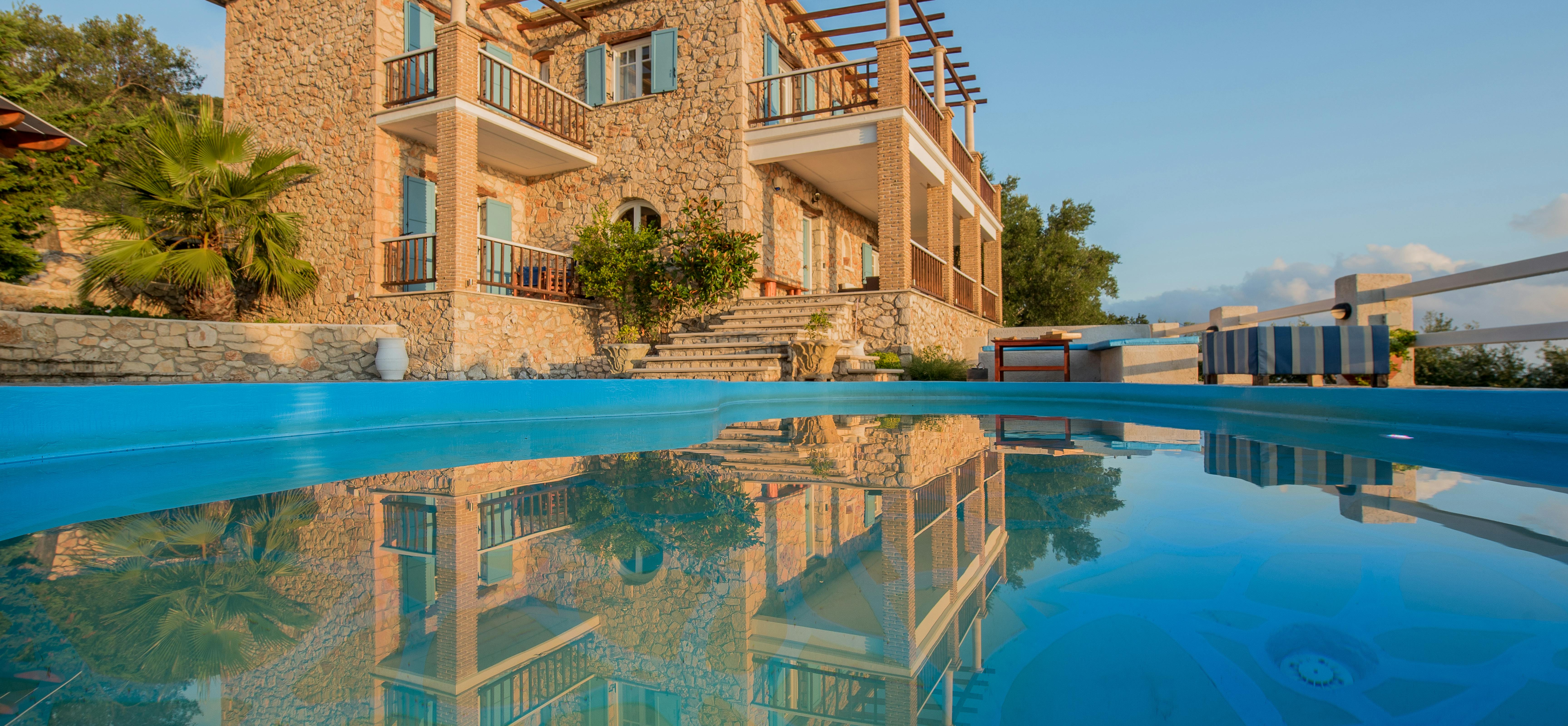 The stone villa reflects perfectly in the still swimming pool water at sunset, creating a mirror image of the Mediterranean-style architecture.