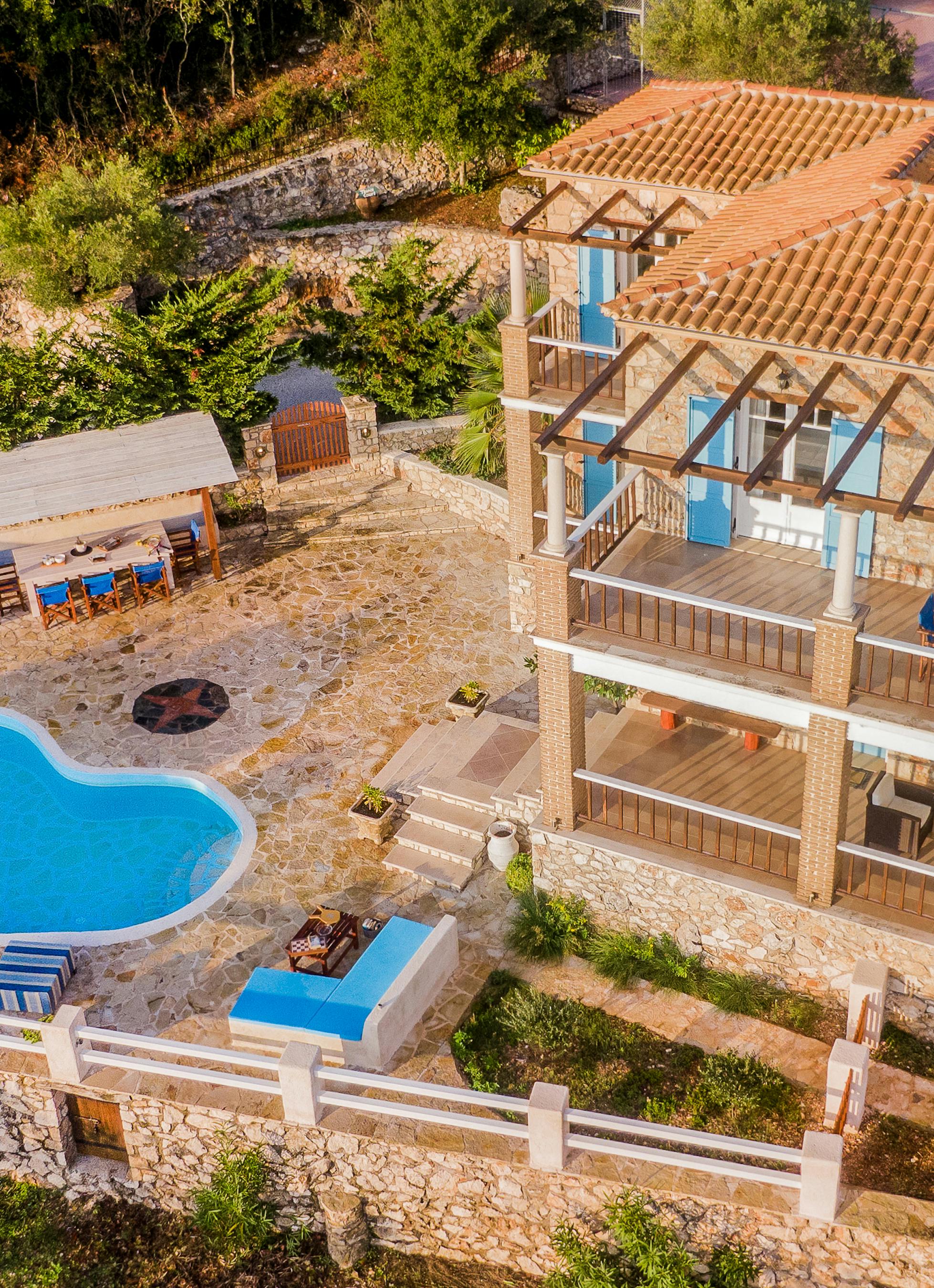 Aerial view of a two-story stone villa with terracotta roof and wooden pergola structures, featuring multiple balconies with blue accents, a kidney-shaped pool, and an outdoor dining area on a terraced hillside property.