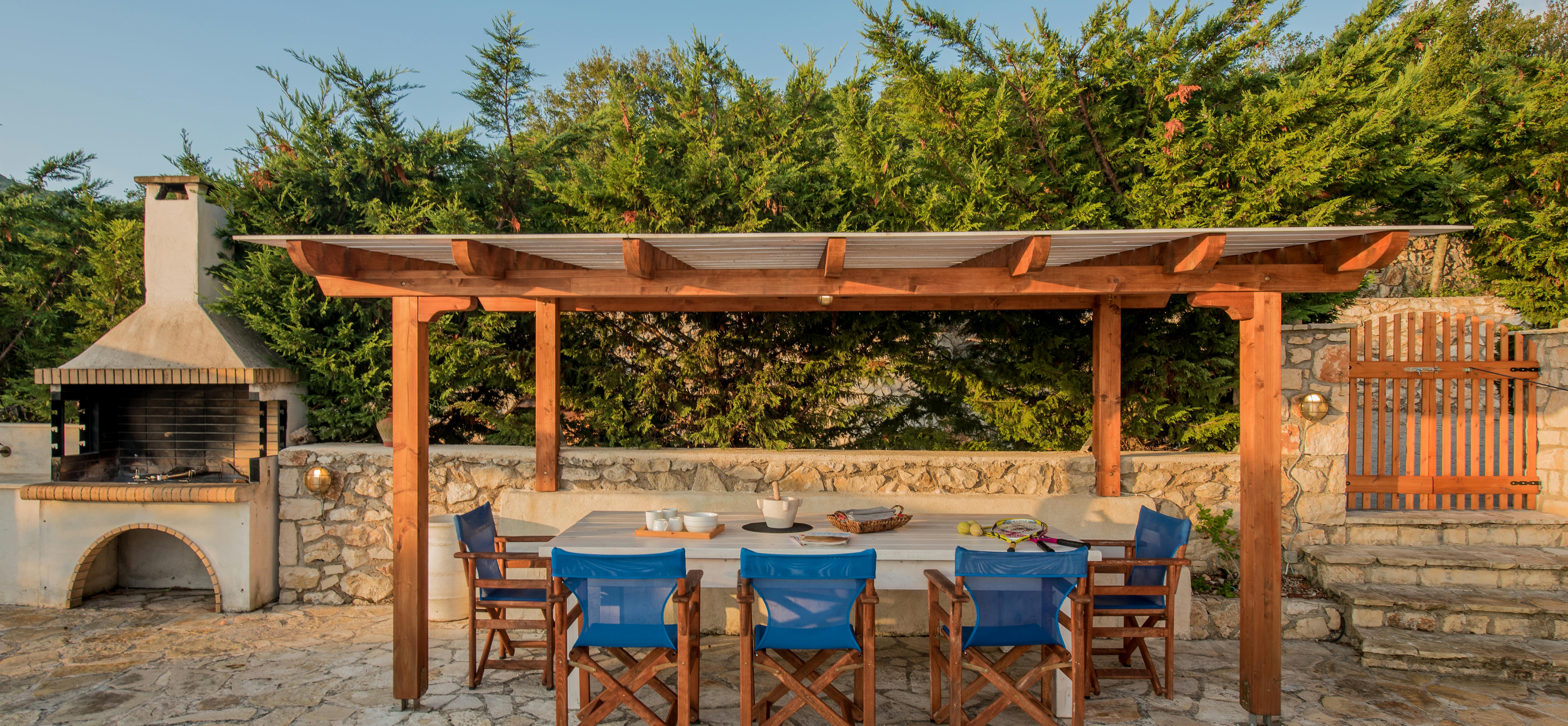 An outdoor dining area under a wooden pergola includes a stone table with blue director's chairs and a built-in barbecue station against a stone wall.