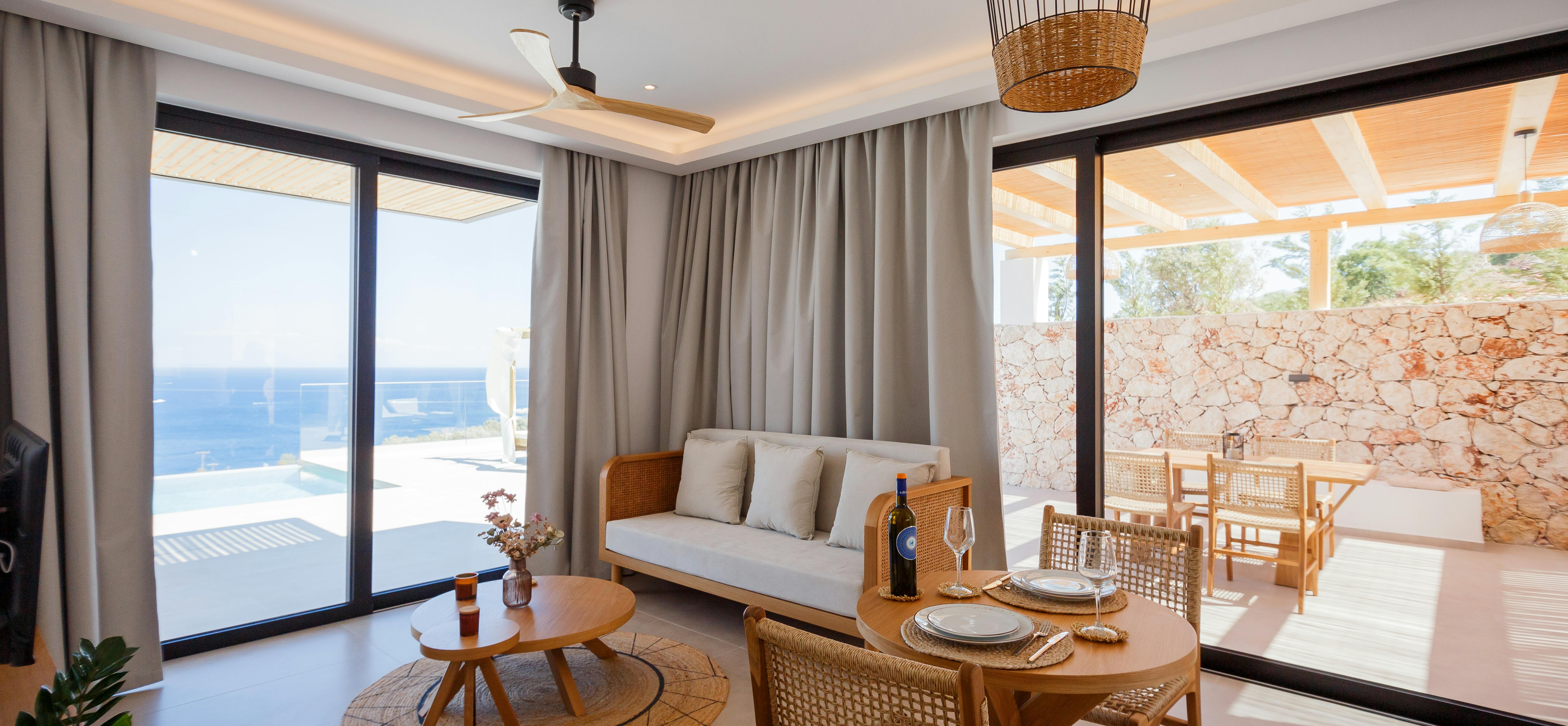 A modern open-plan living and dining area features natural wood furniture with woven details, floor-to-ceiling windows opening to a terrace with ocean views, and a cohesive neutral palette with natural textures.