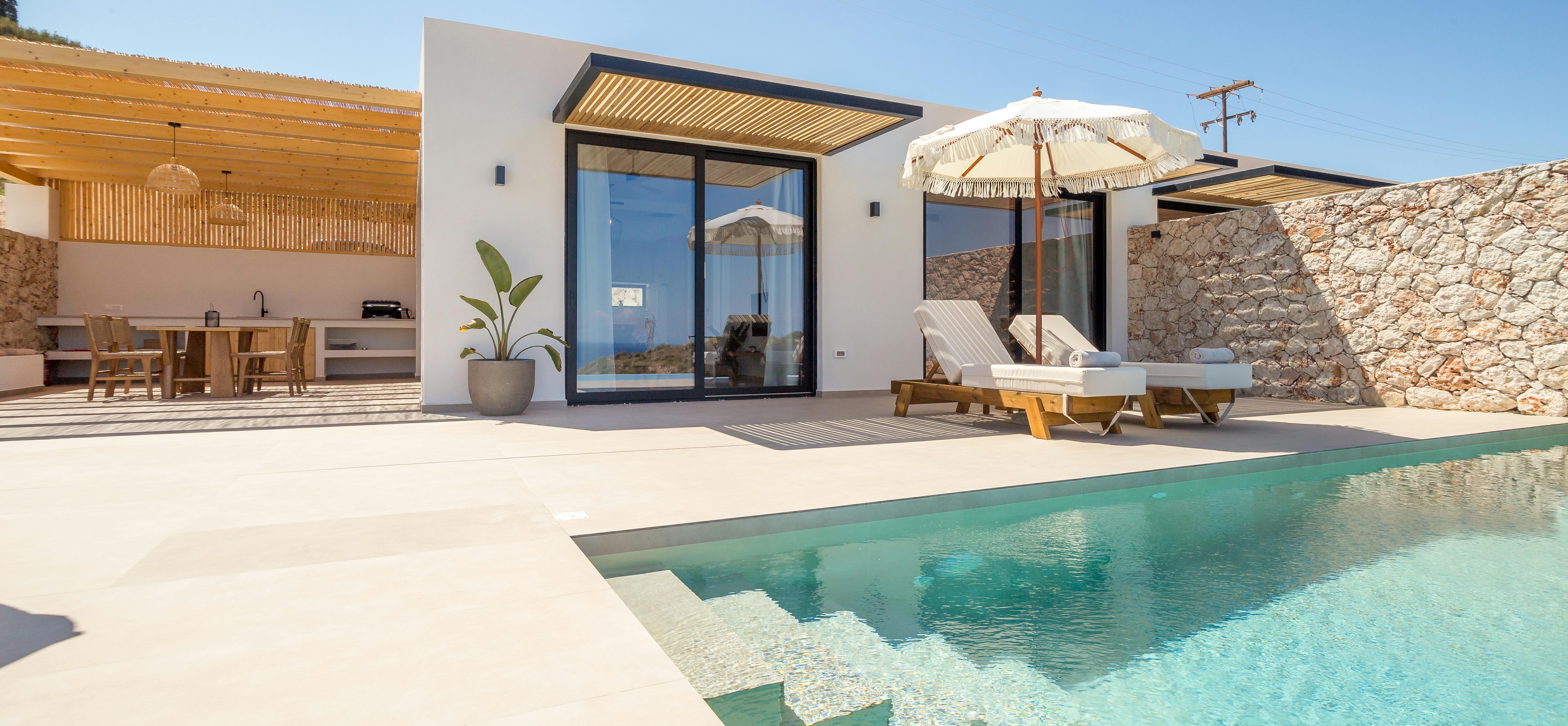 A contemporary white villa exterior showcases floor-to-ceiling glass doors, wooden slatted pergola details, and a turquoise infinity pool with integrated steps, flanked by stone walls and sun loungers under fringed umbrellas.