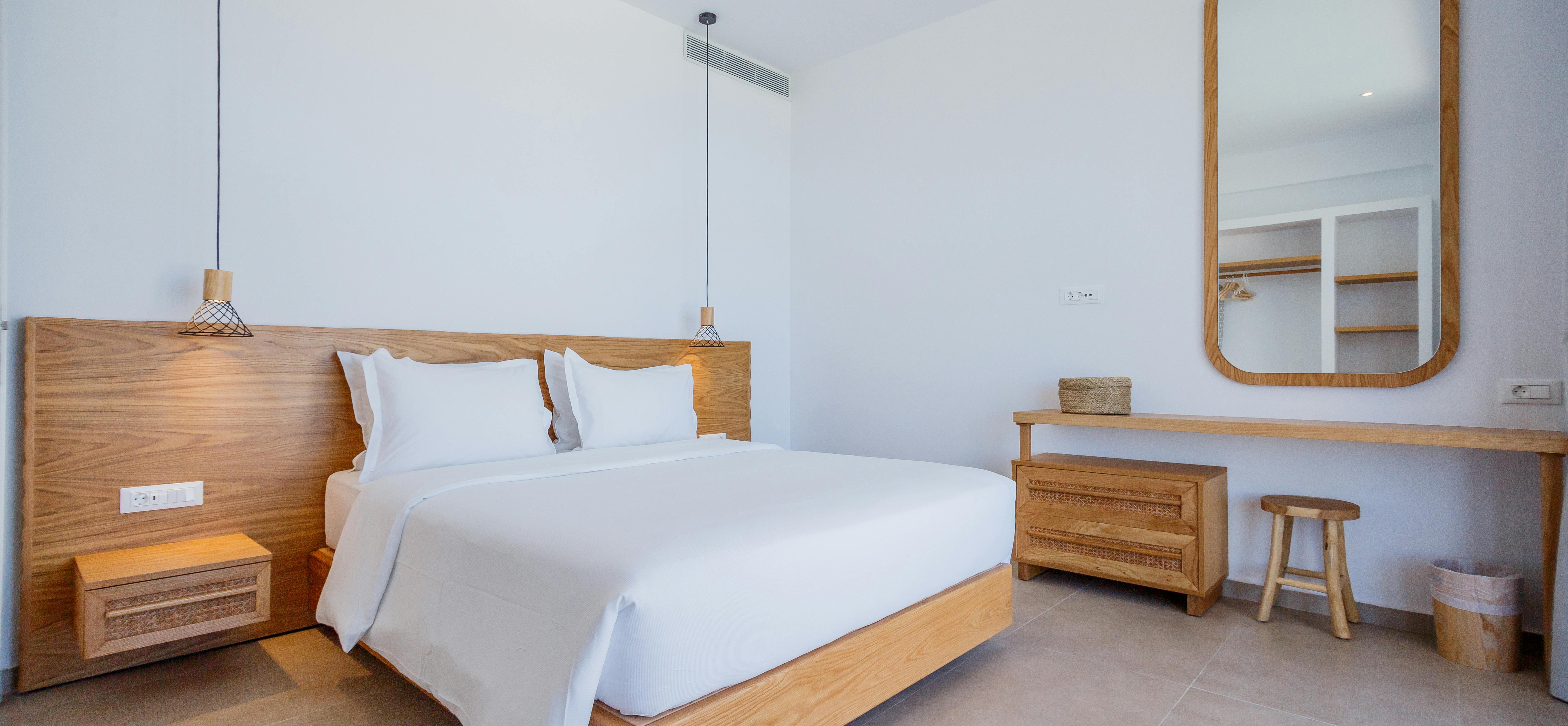 A minimalist bedroom with white linens on a wooden platform bed features a rounded mirror above a floating desk with stool, pendant lighting, and natural wood accents throughout.
