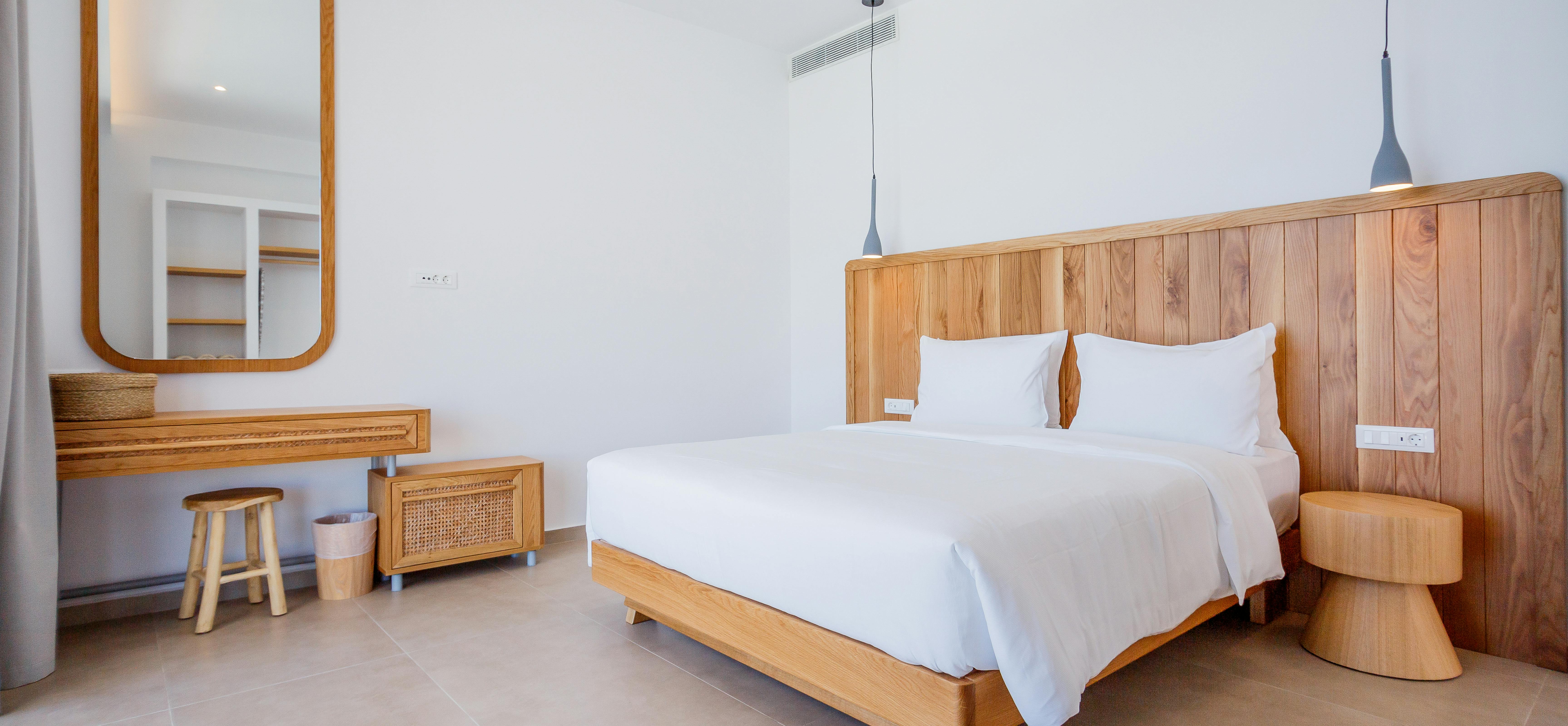 A spacious bedroom with tall wooden headboard, white linens, pendant lights, and rounded mirror includes a floating desk area and built-in wardrobe in a neutral palette.