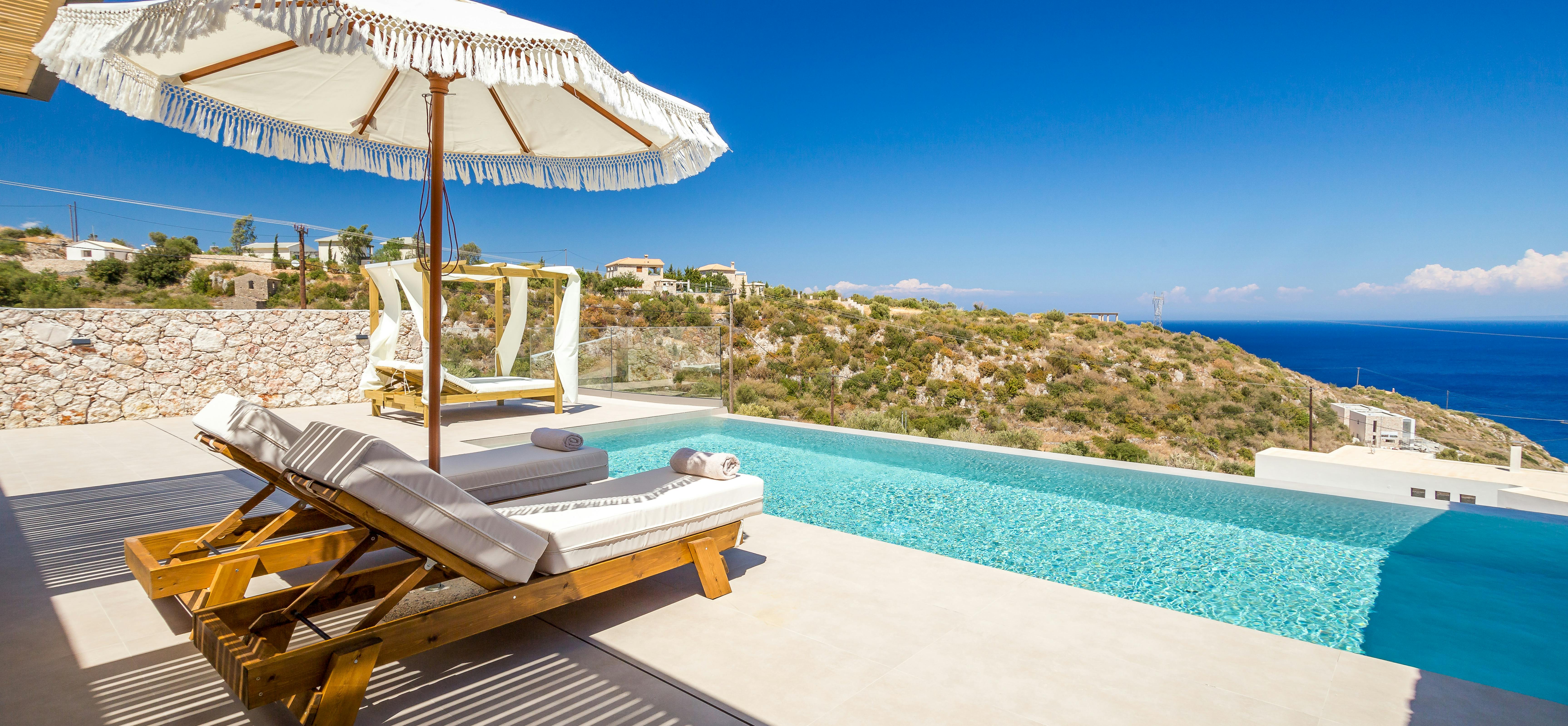 A hillside pool terrace with wooden sun lounger and fringed umbrella overlooks dramatic coastal views, featuring turquoise water and a modern white villa with stone walls in the background.