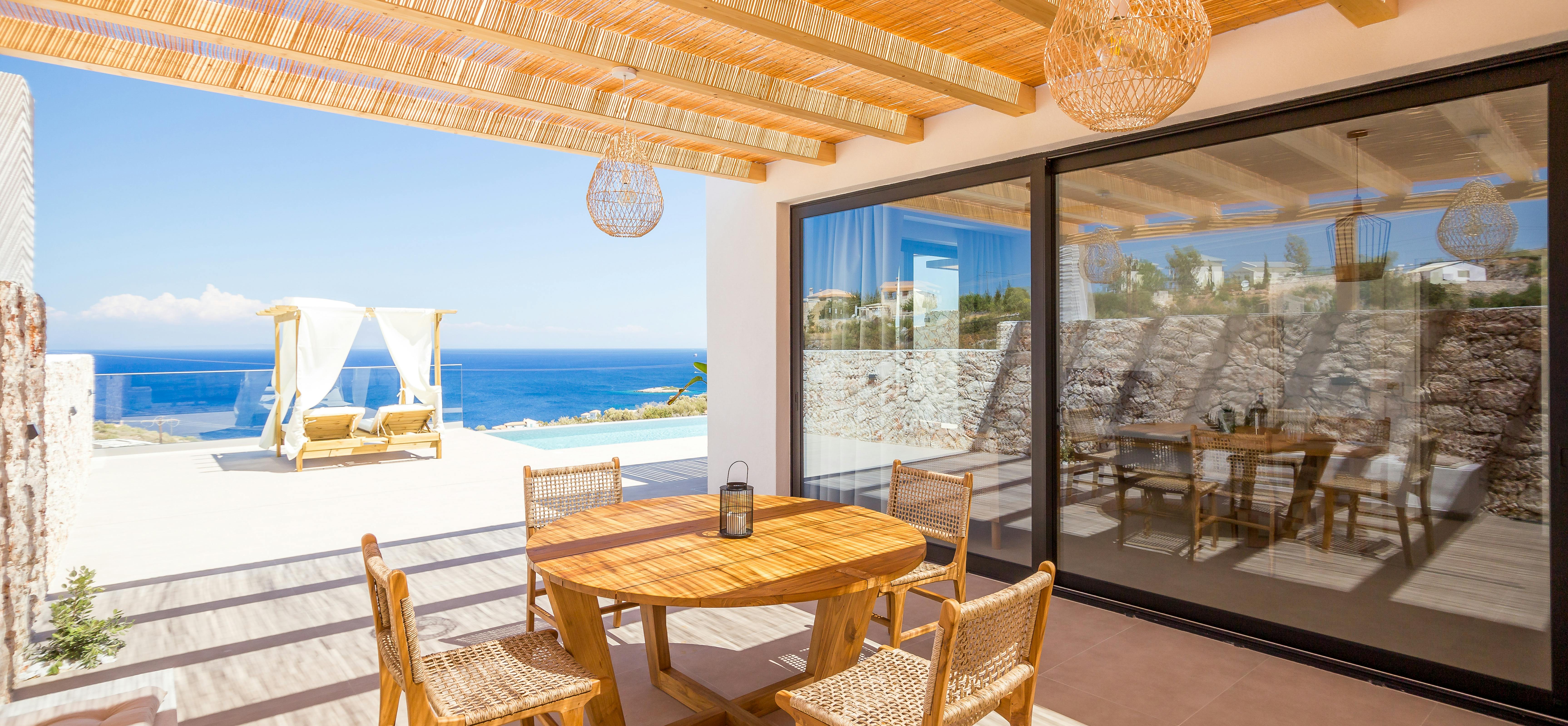 A covered dining terrace with bamboo ceiling features a round wooden table with woven chairs, wicker pendant lights, and frames views of a pool, daybed, and expansive sea vista beyond stone walls.