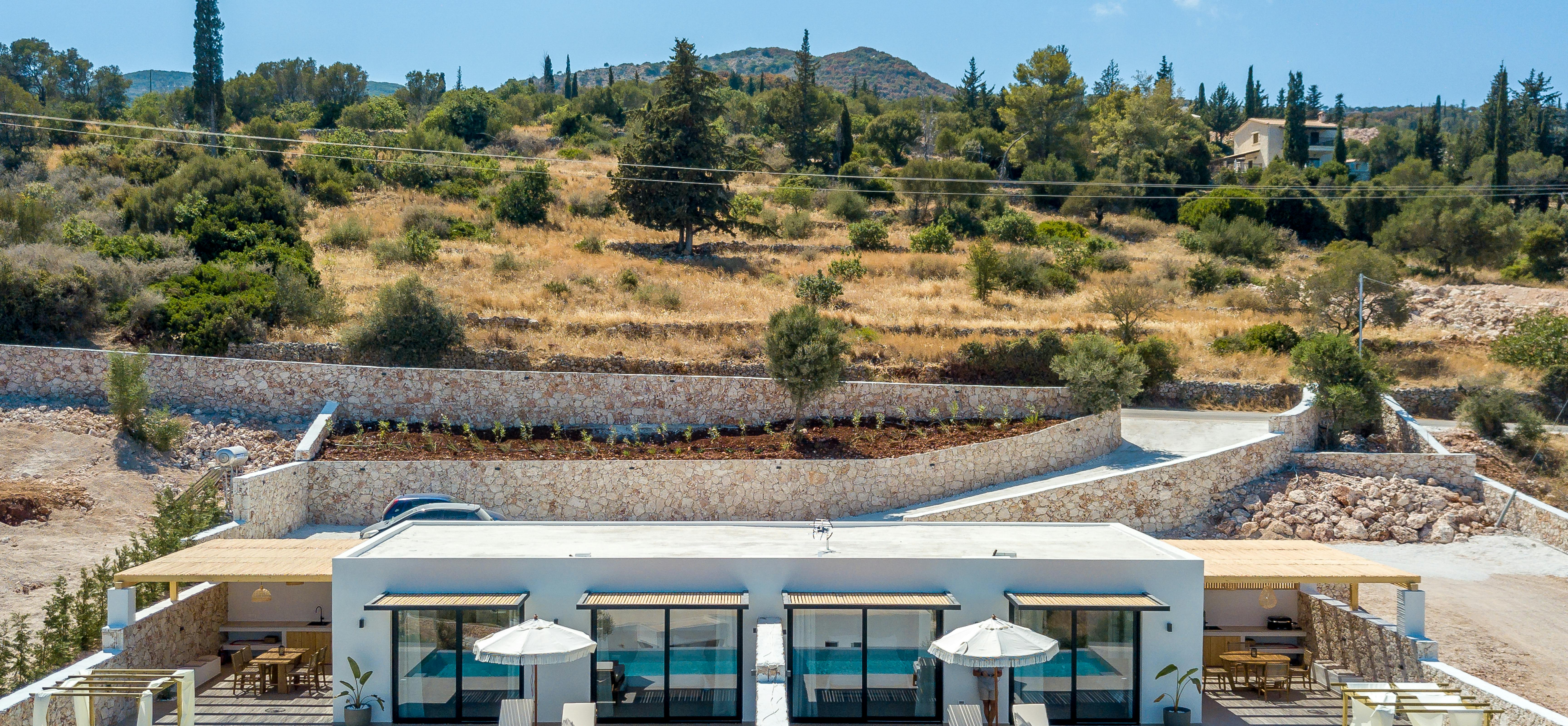 Frontal view of modern white villa complex with multiple units featuring floor-to-ceiling windows, wooden pergola roofs, shared infinity pools, and terraced landscaping against a hillside backdrop.