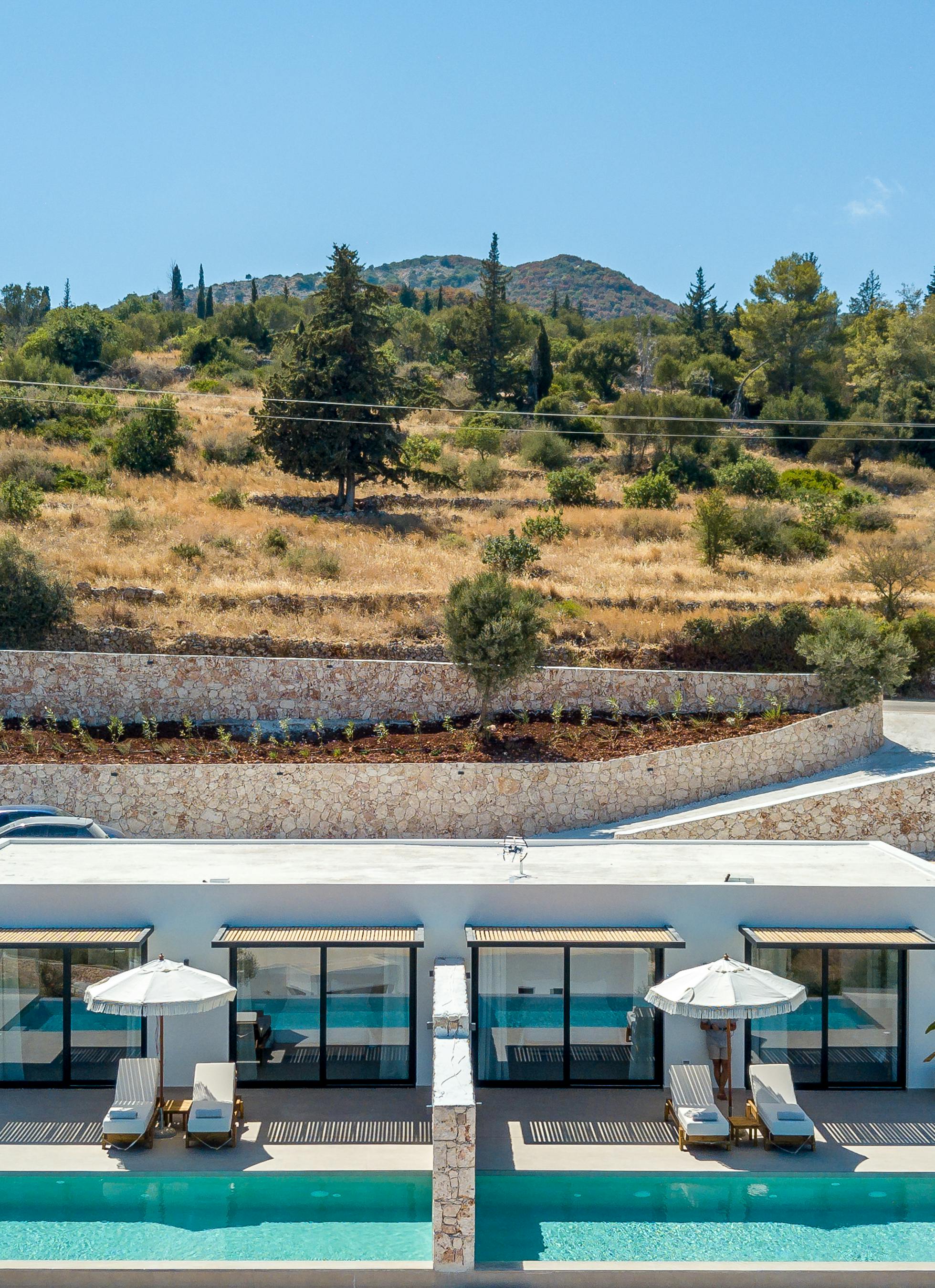 Frontal view of modern white villa complex with multiple units featuring floor-to-ceiling windows, wooden pergola roofs, shared infinity pools, and terraced landscaping against a hillside backdrop.