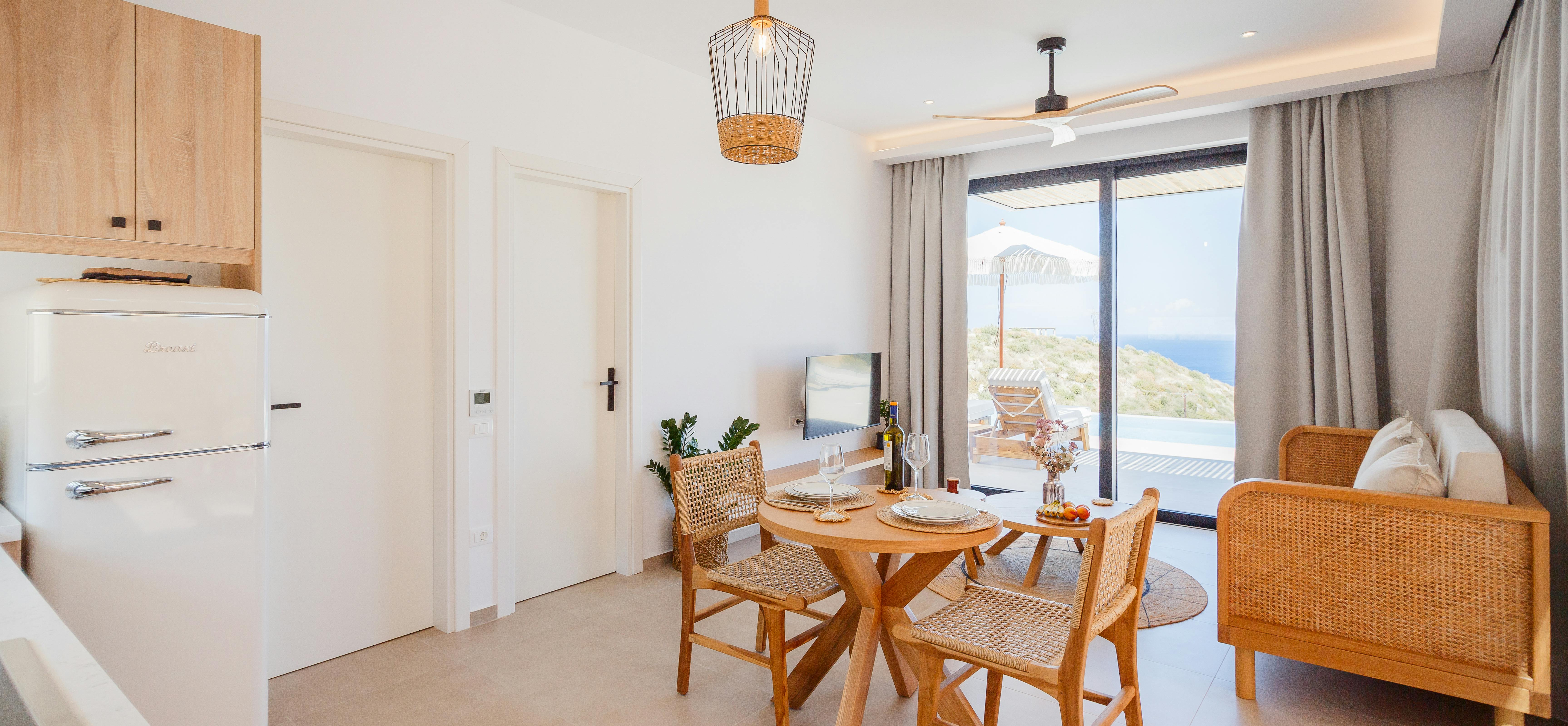 A light-filled living and dining space features round wooden table with woven chairs, modern kitchenette with vintage-style refrigerator, and terrace access with coastal panoramas beyond grey curtains.