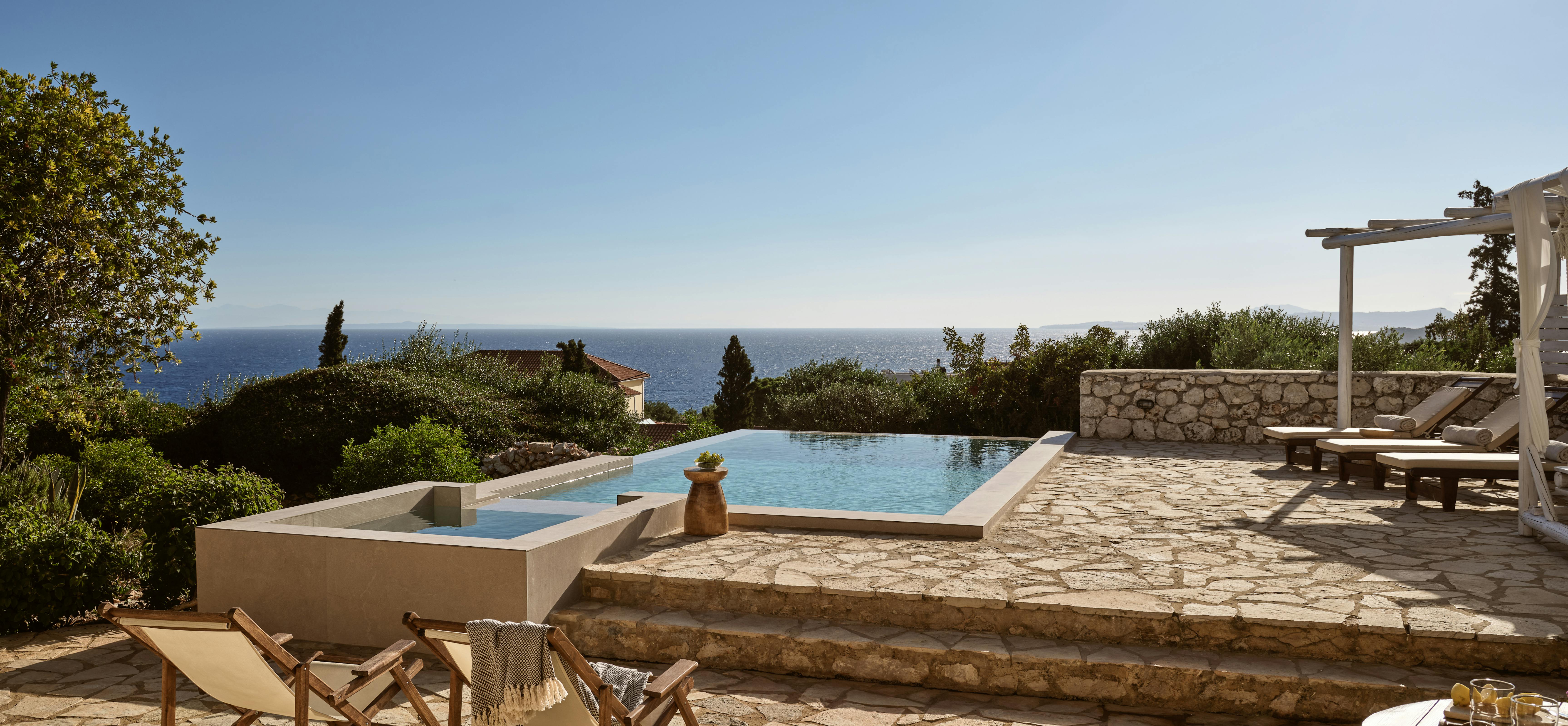 Pool overlooking the sea with stone terrace, wooden sun loungers, and white pergola providing shade beside Mediterranean vegetation.