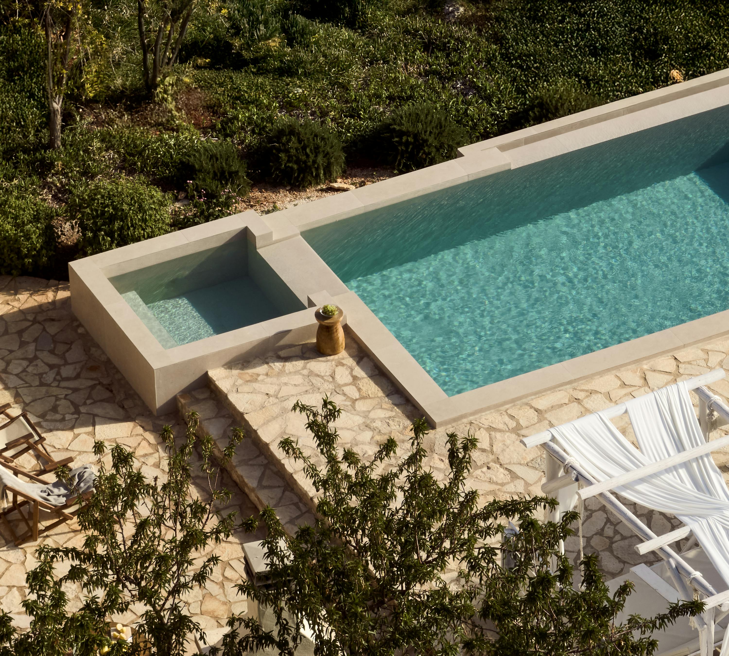 Aerial view of minimalist pool with integrated spa, surrounded by irregular stone paving and white sun loungers beneath trees.