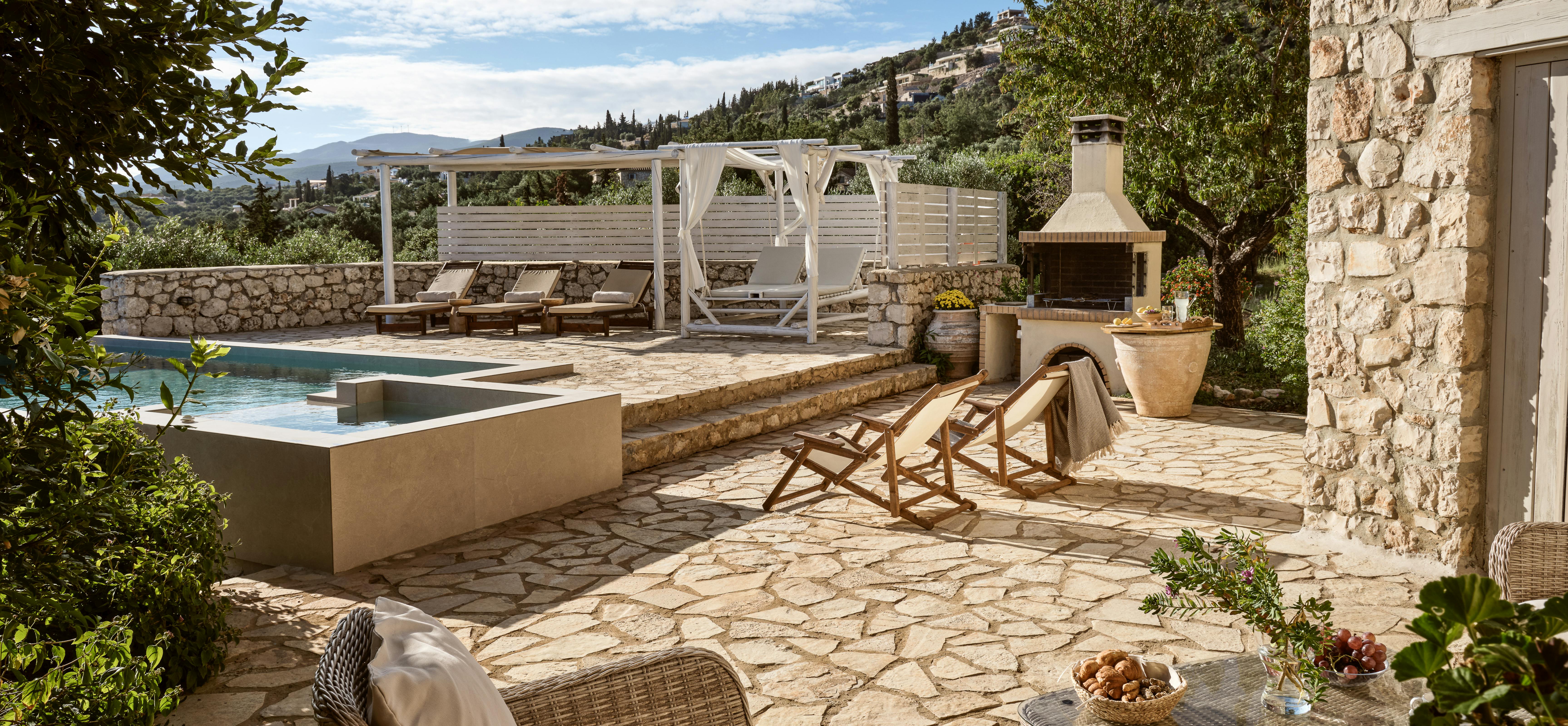 Terraced outdoor space featuring pool, white pergola with curtains, built-in barbecue, and stone paving with mountain backdrop.