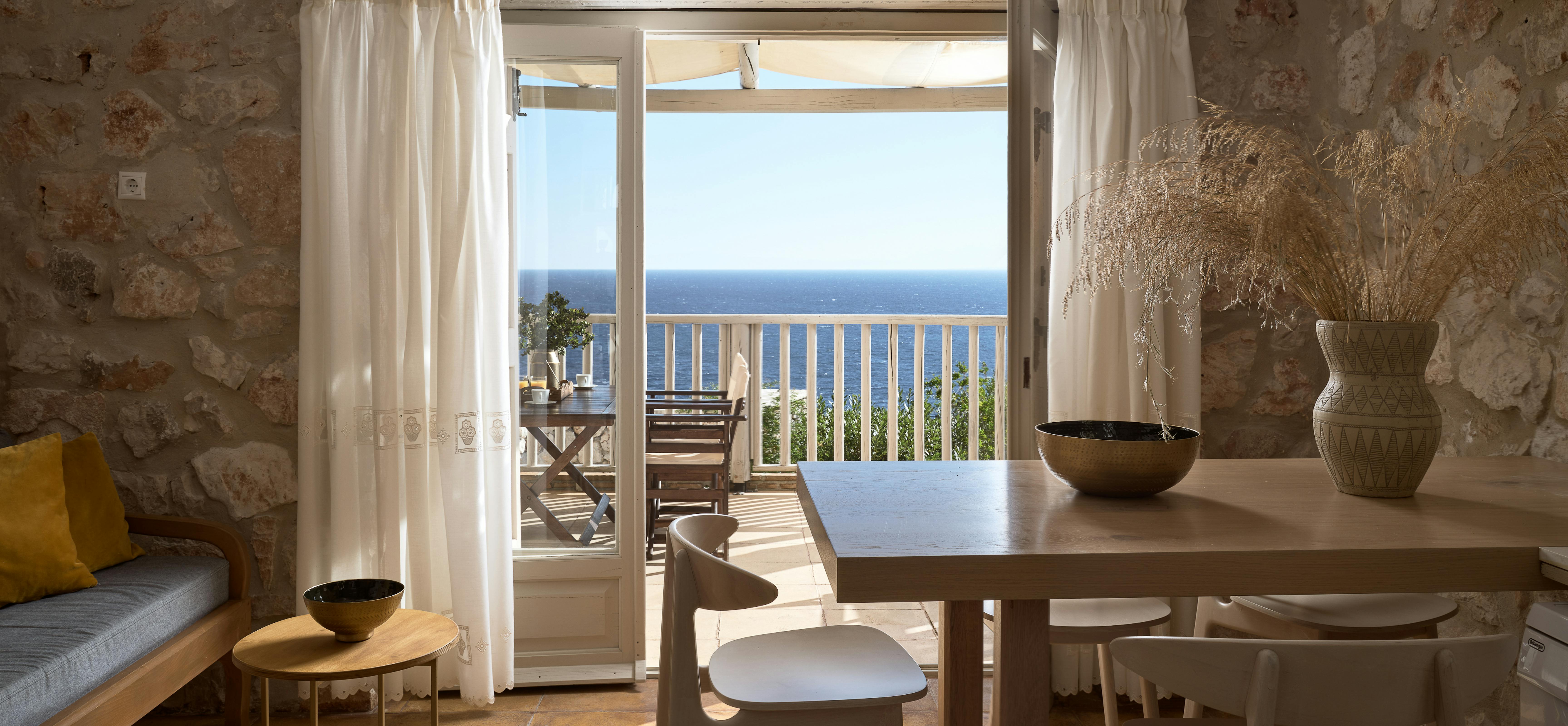 Dining area with stone walls, wooden table and chairs, crystal chandelier, and French doors opening to balcony with ocean panorama.