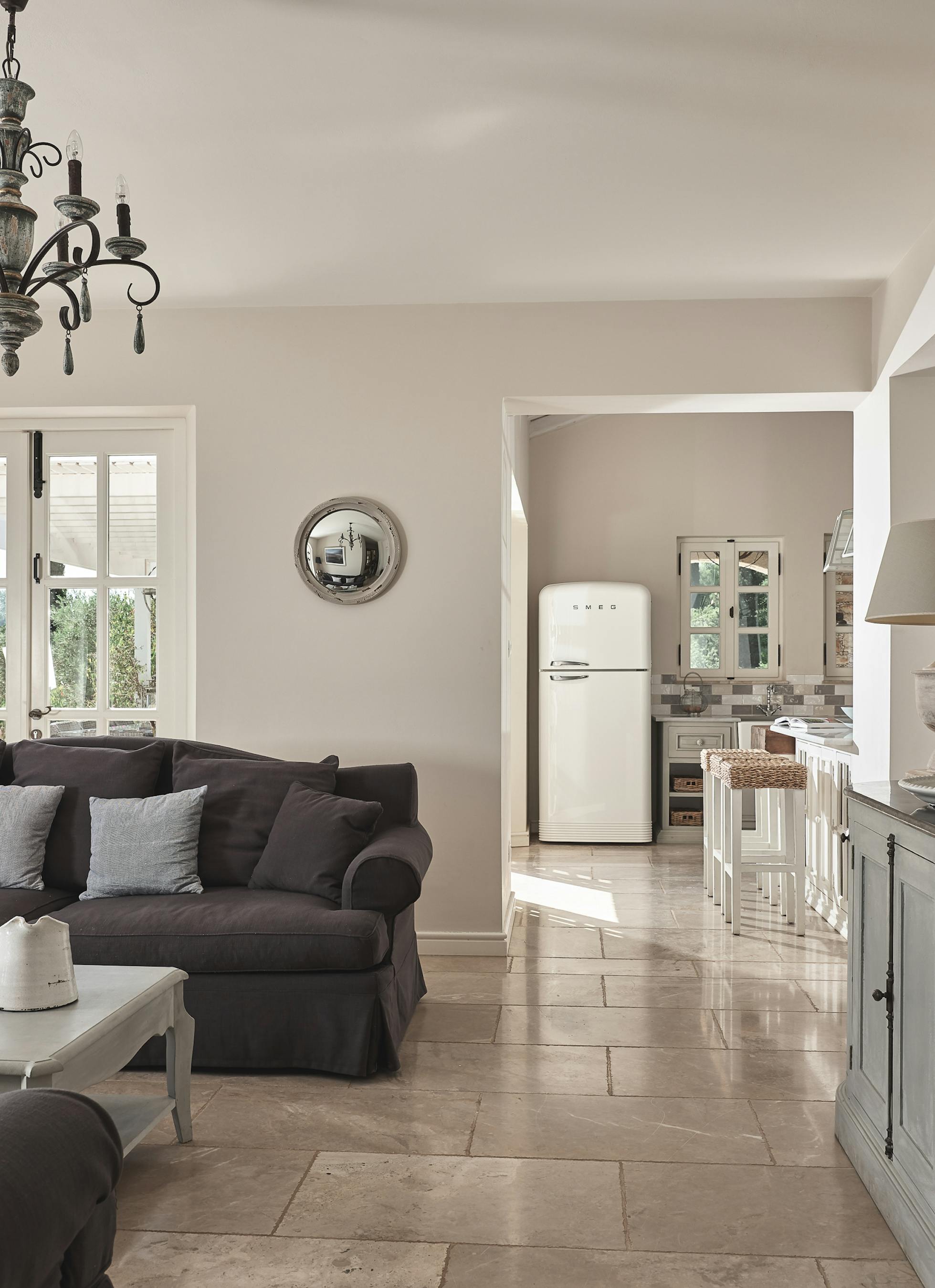 Open-plan living area with chandelier and dark sofas, connecting to bright farmhouse kitchen through white doorway.