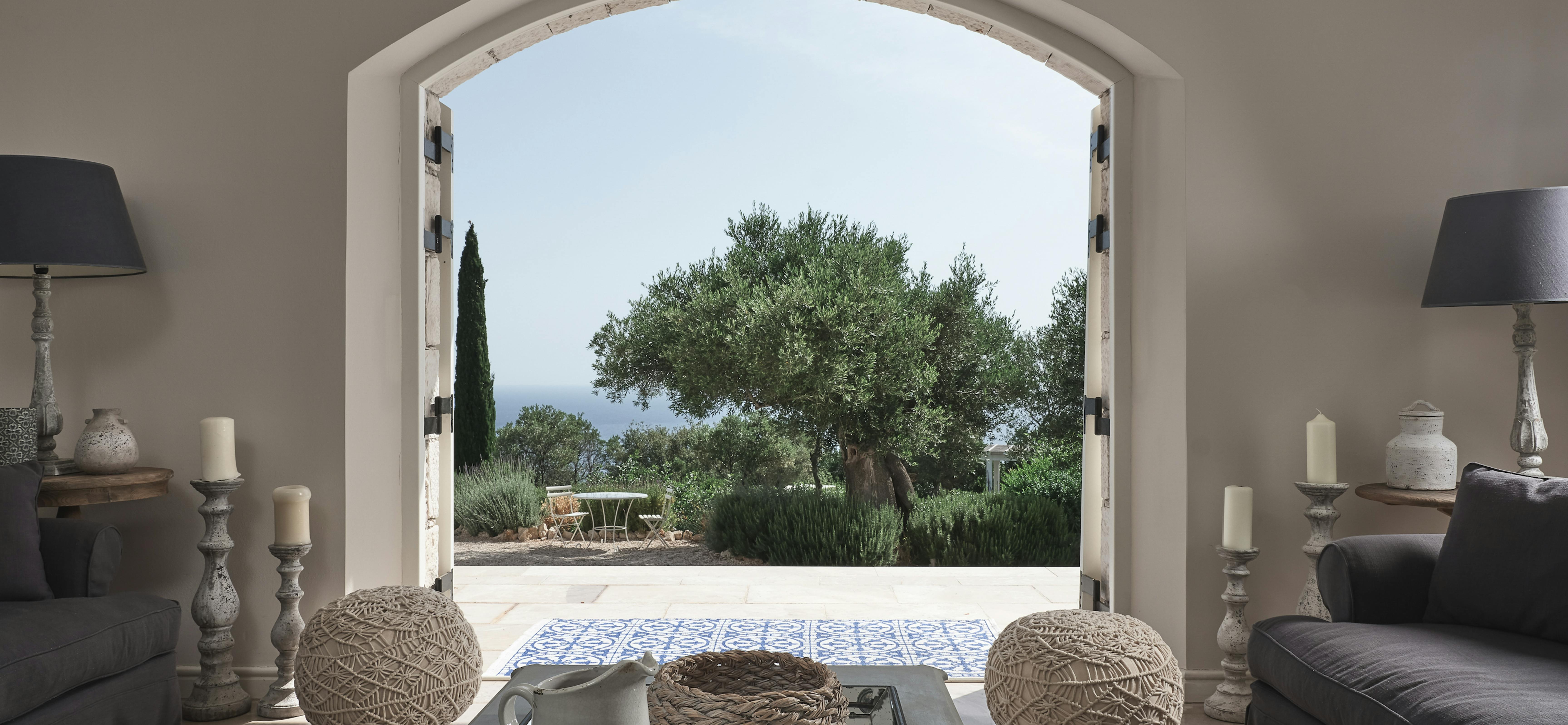 View through arched doorway to terrace with olive tree and coastal vista, framed by neutral-toned living space.