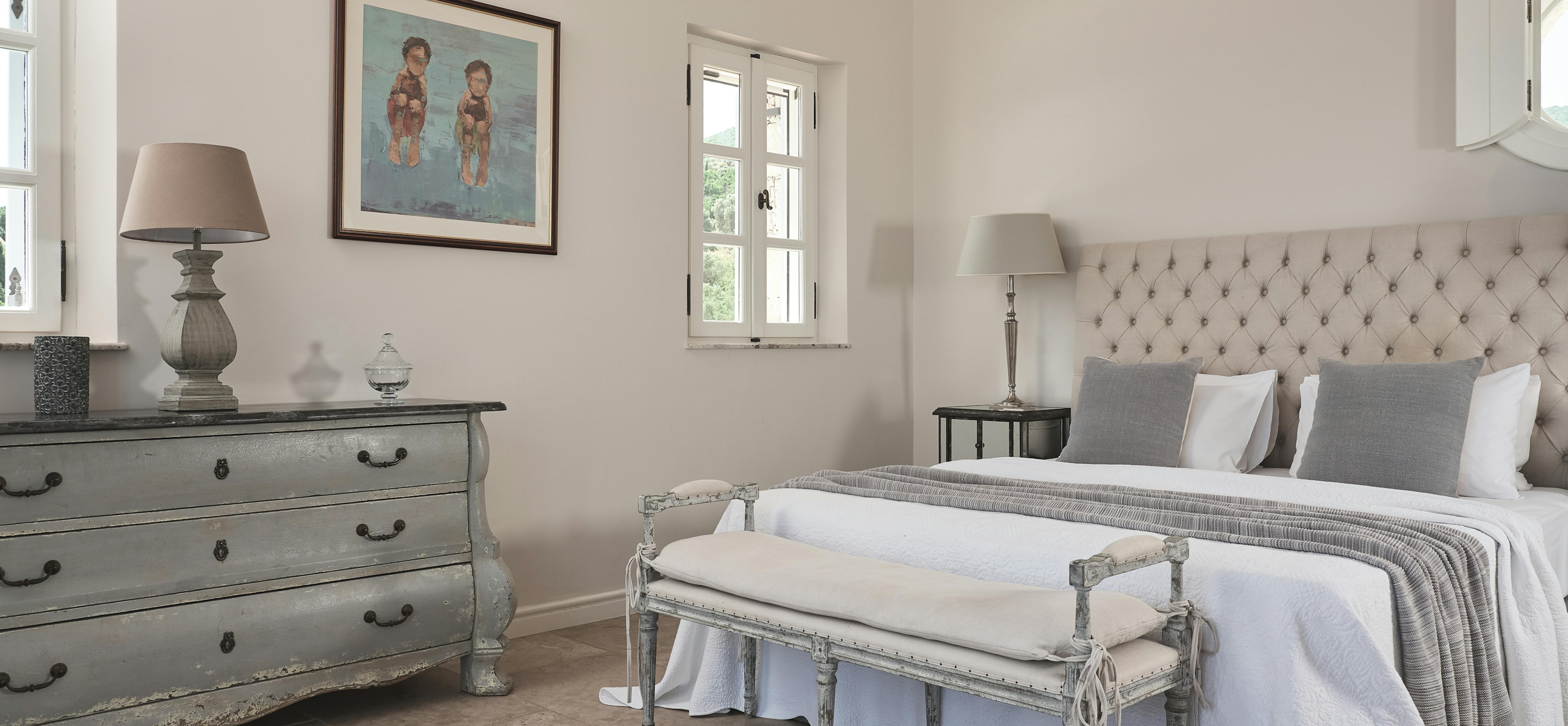 Serene bedroom with tufted headboard and distressed furniture, featuring exposed beam ceiling and French doors.