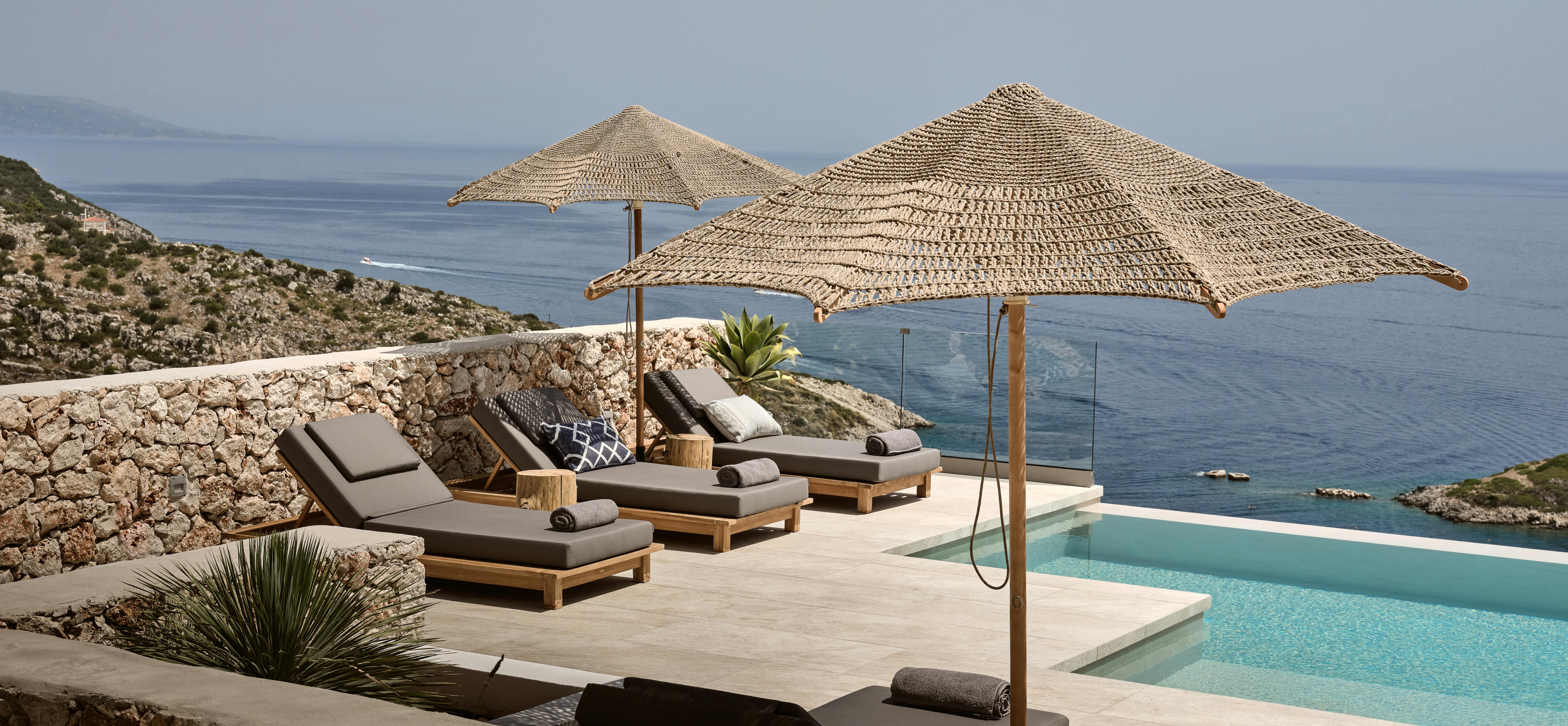 Infinity pool terrace with wooden loungers, woven umbrellas, and dramatic coastal views. Multi-level stone terracing overlooks turquoise Mediterranean waters.