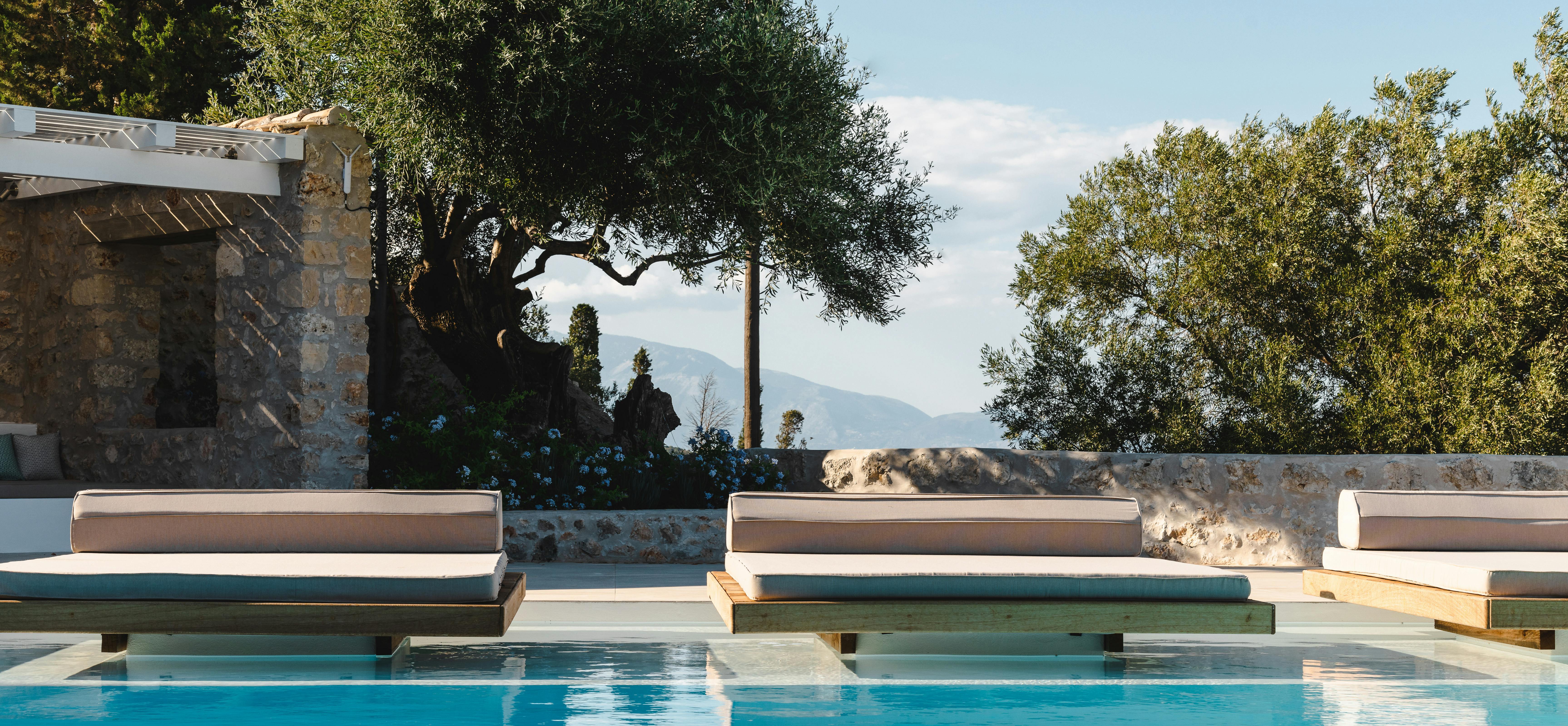 In-pool loungers with ocean views, mature olive trees, rustic stone walls, and distant mountains framing the Mediterranean Sea.