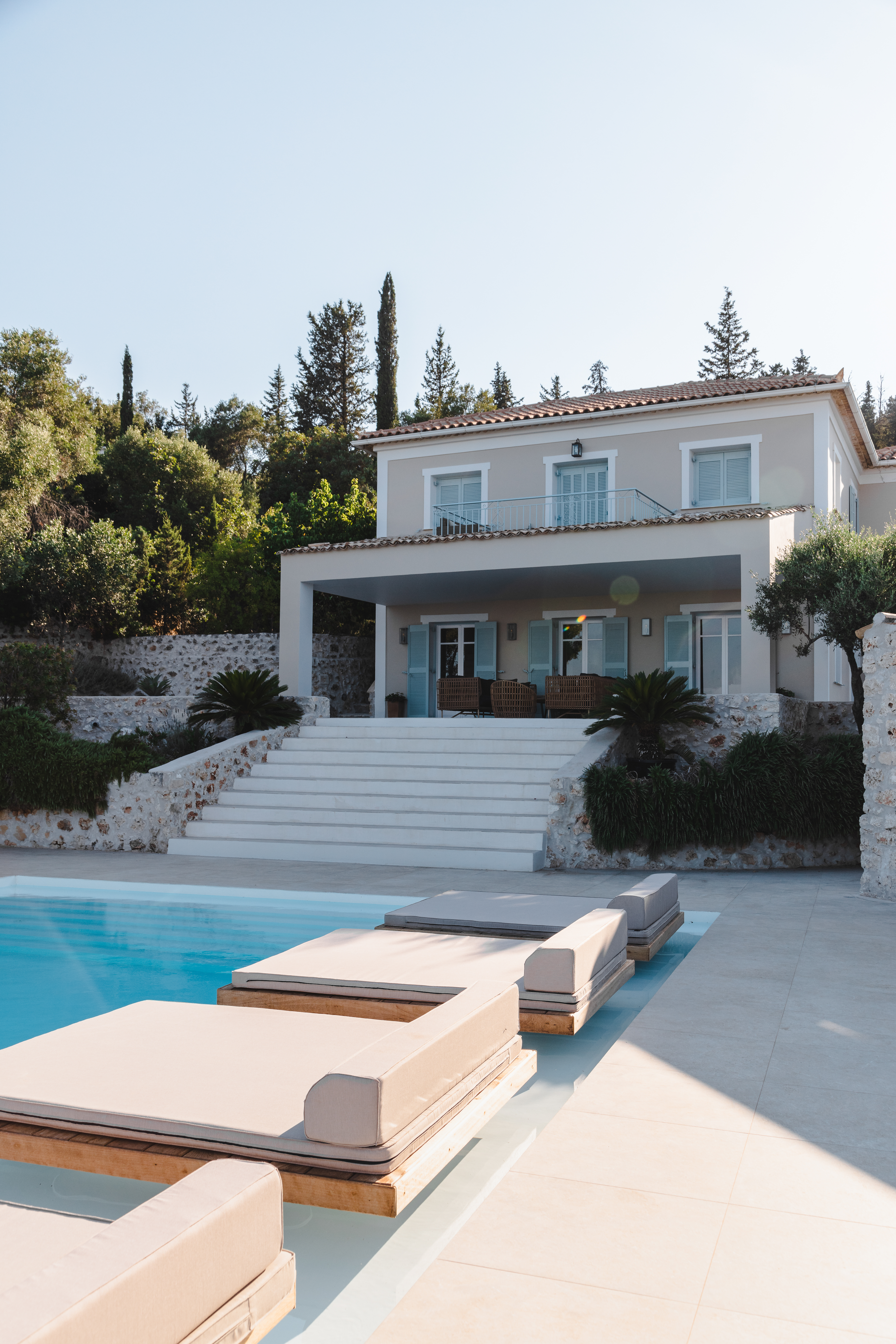 Pool deck view showing two-story white villa with blue shutters, grand staircase, stone walls, and modern loungers beside infinity pool.