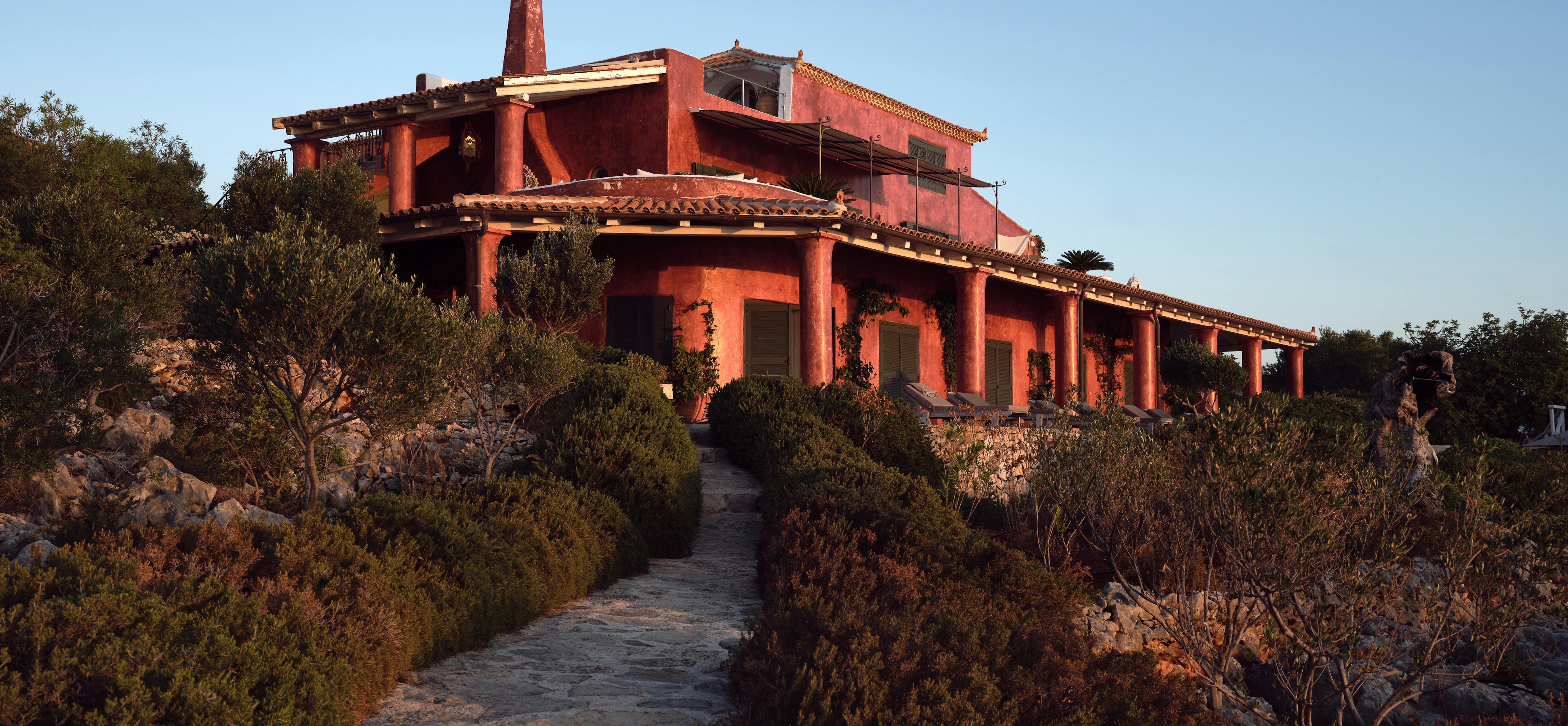 Dramatic terracotta-coloured Mediterranean villa at dusk perched on hillside with multiple levels, pergolas, and winding stone path through native vegetation.
