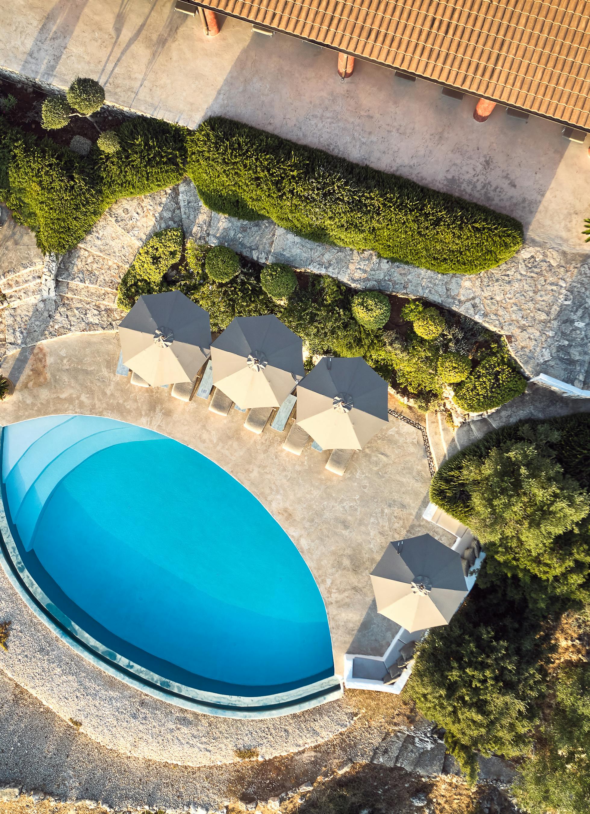 Aerial view of curved infinity pool with beige umbrellas, stone terrace, natural landscaping, and terracotta-roofed villa visible above.
